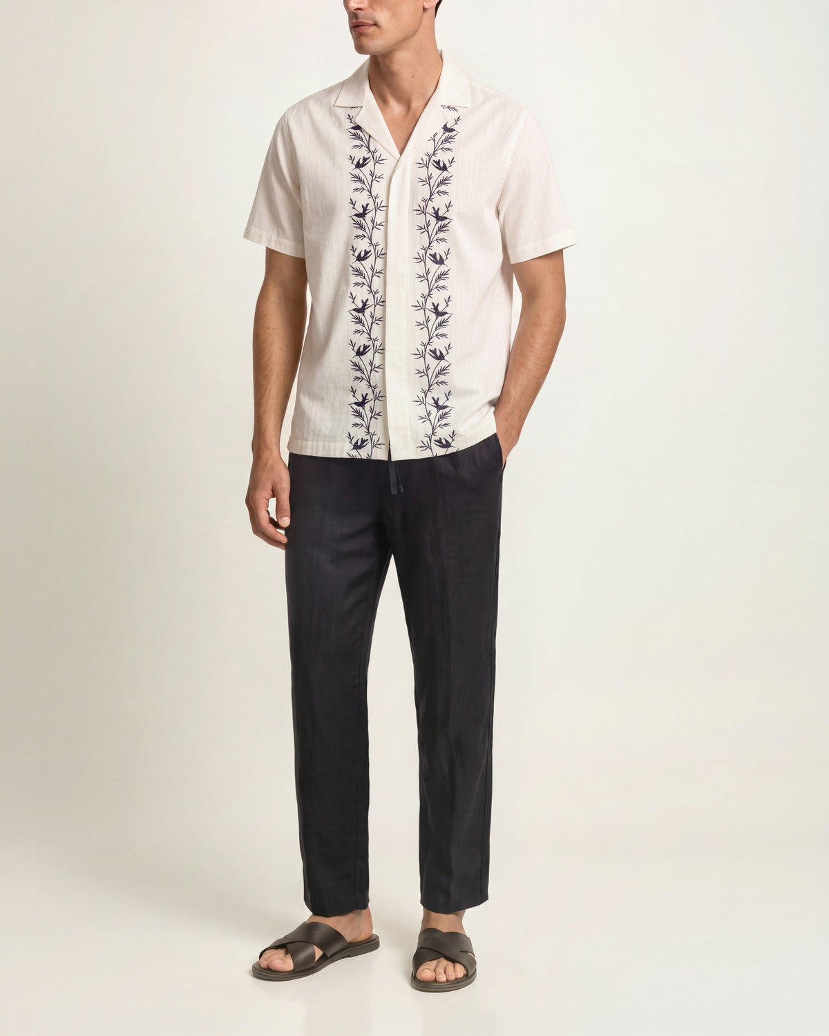 Man wearing white short sleeve shirt with black embroidery detail and black trousers and brown sandals on a plain background