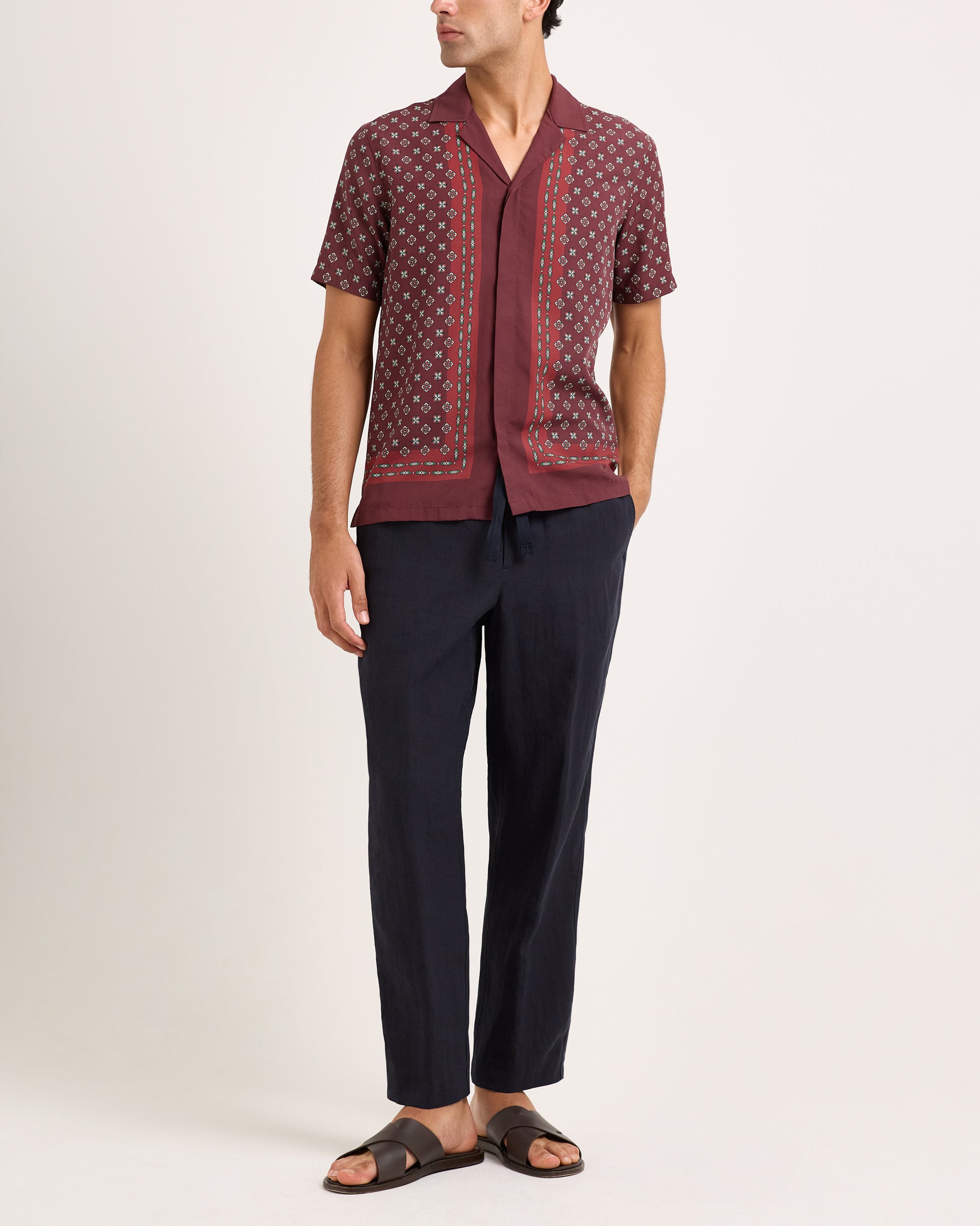 Model wearing maroon short-sleeve shirt with geometric print and navy trousers on a white background