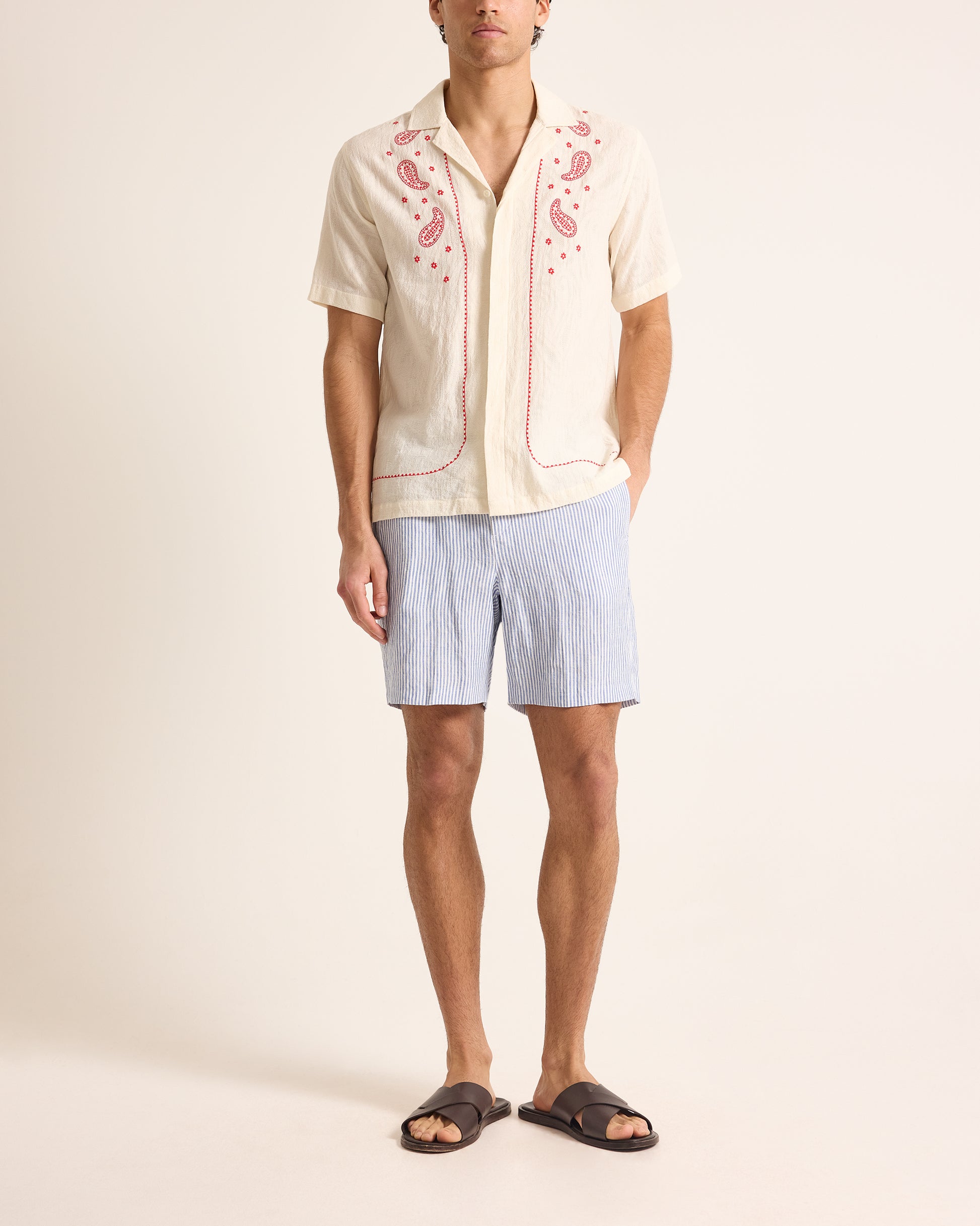 Man wearing a cream embroidered shirt and light blue shorts on a plain background