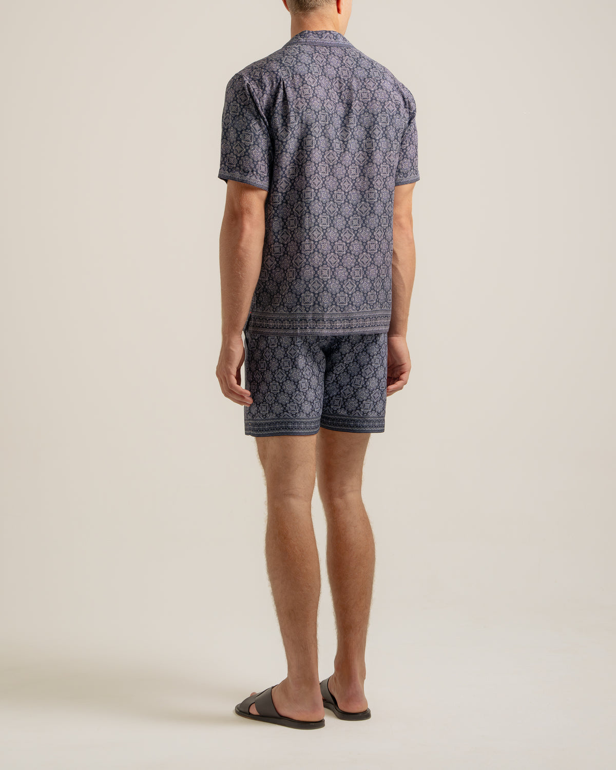 A man viewed from the back wearing a matching short-sleeve button-up shirt and shorts set in a navy blue and beige geometric pattern, paired with dark brown slide sandals, against a solid light beige studio background.