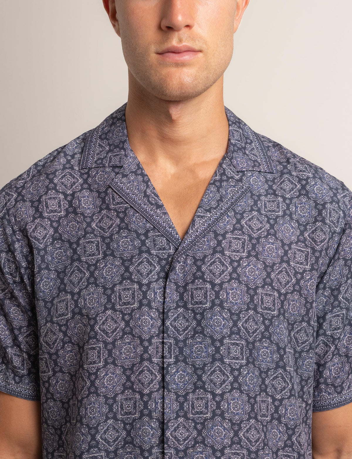 A man wearing a short-sleeve button-front camp shirt with a blue and gray geometric medallion pattern, shown from the chest up against a plain light beige background.