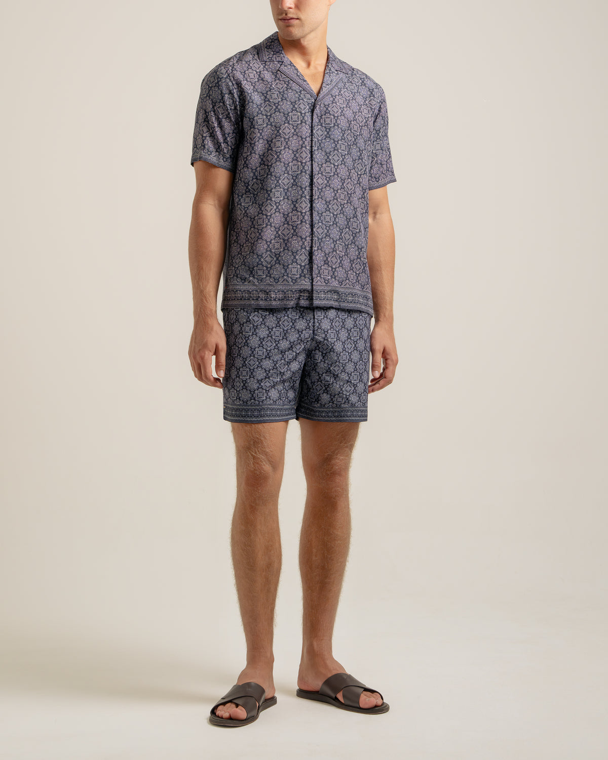 A man modeling a matching short-sleeve button-front camp shirt and shorts set in a dark navy blue and light beige geometric pattern, paired with dark brown slide sandals, against a plain light beige studio background.