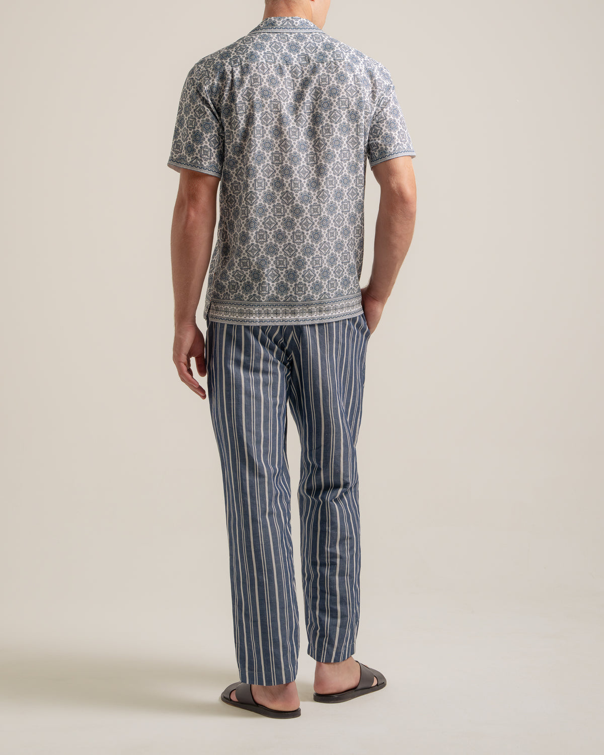 A man viewed from the back wearing a short-sleeve patterned t-shirt and blue-and-white vertically striped pajama pants, standing against a plain light beige studio background.