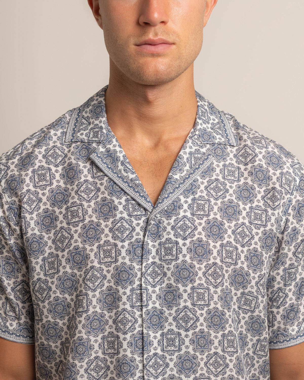 A close-up of a man wearing a short-sleeve button-up shirt with a blue and gray geometric pattern on a white background, set against a plain beige studio backdrop.