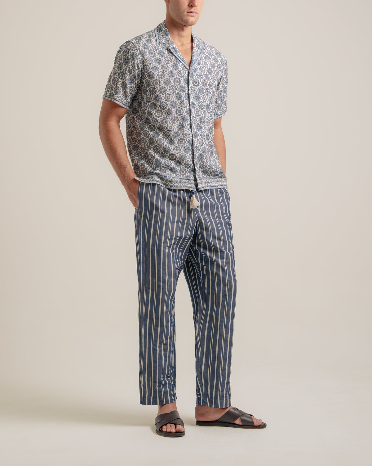A man modeling a matching two-piece pajama set consisting of a short-sleeve button-up shirt with a blue and white geometric pattern and striped pants with a drawstring waist, paired with black slide sandals, against a solid light beige background.