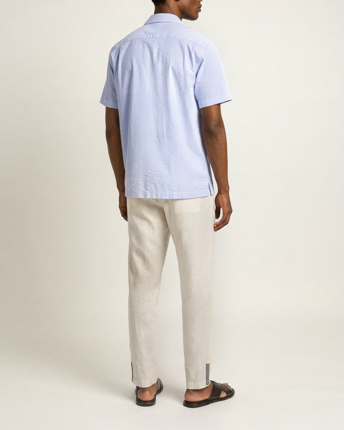 Man viewed from the back wearing blue short sleeve shirt with beige trousers and brown sandals on a plain background