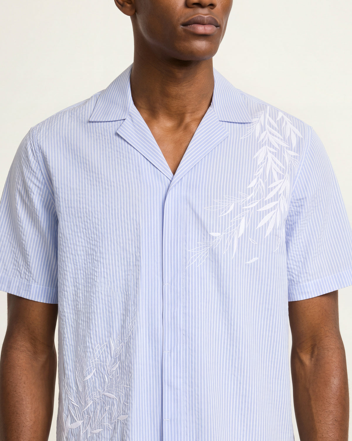 Close-up of a man wearing a blue striped seersucker shirt featuring a white embroidery detail on a white background
