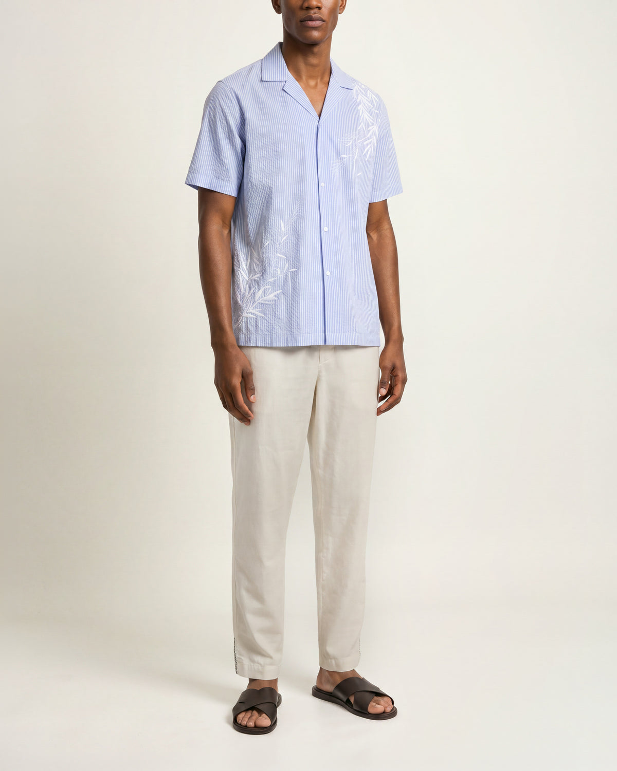 Man wearing blue short sleeve shirt with beige trousers and brown sandals on a plain background