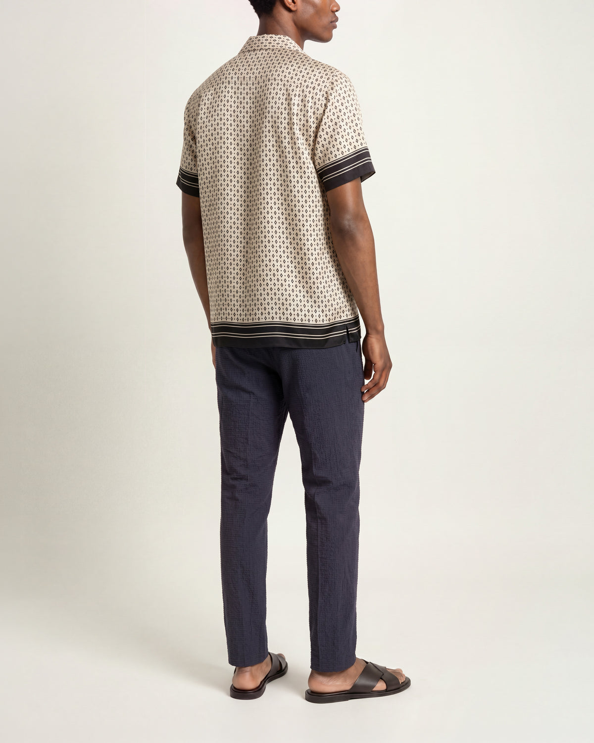 Back view of a man wearing a beige patterned silk shirt with short sleeves and dark navy trim with navy trousers and brown sandals on a plain background.