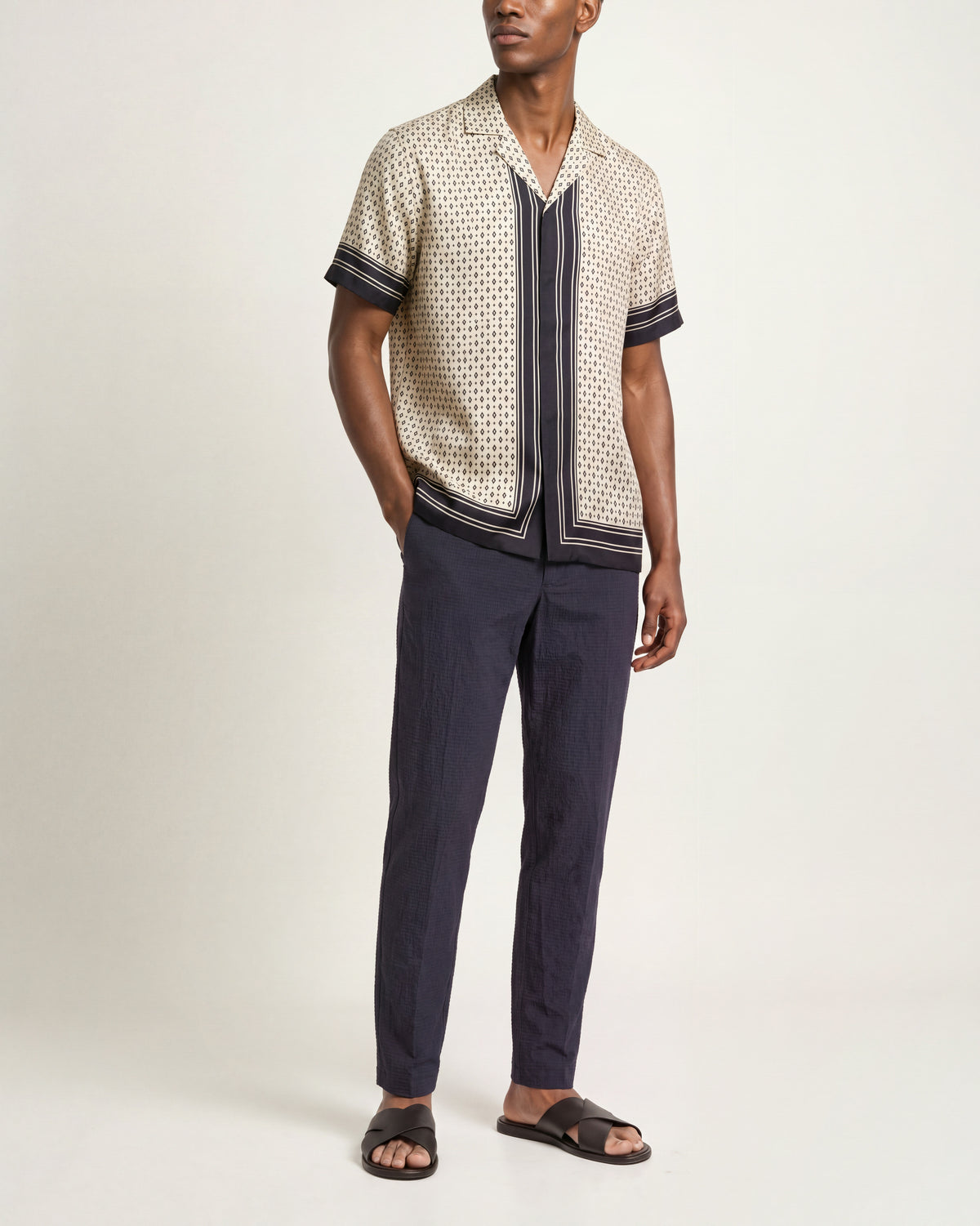 Man wearing a beige patterned silk shirt with short sleeves and dark navy trim with navy trousers and brown sandals on a plain background.