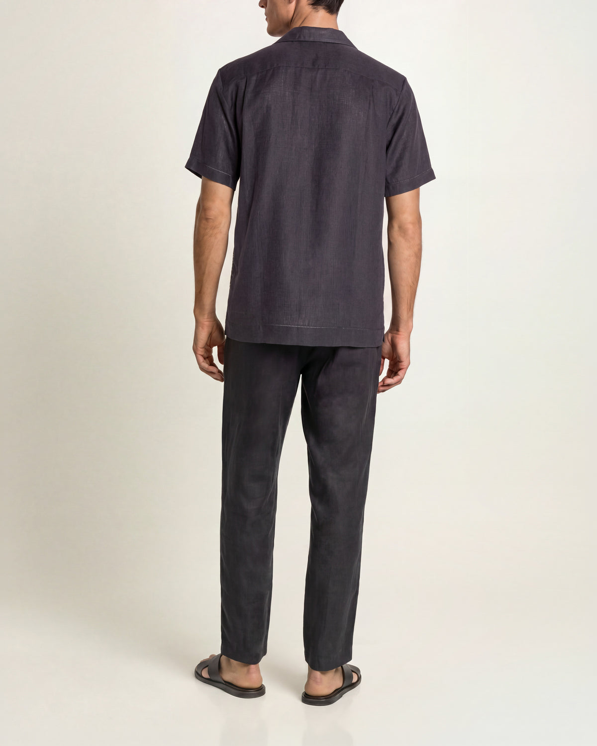A man viewed from the back wearing a dark charcoal navy short-sleeve button-up shirt and matching dark trousers, paired with black sandals, against a plain studio background.