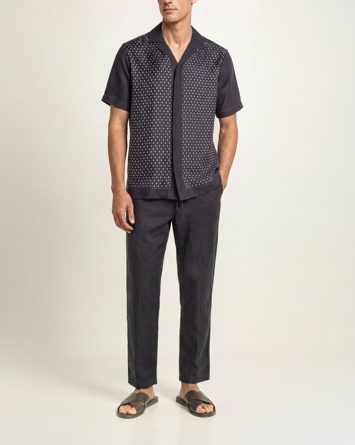 A model wearing a dark navy short-sleeve button-up shirt with a white geometric pattern on the front panel, paired with dark charcoal pants and brown slide sandals, against a plain studio background.