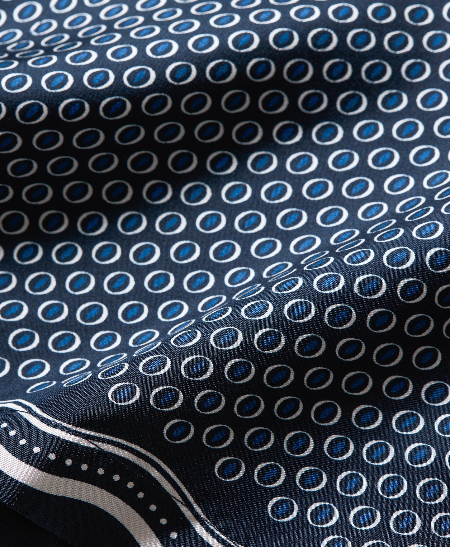 A close-up of a patterned fabric with a repeating blue and white oval motif on a dark navy background, with a decorative white border visible in the lower left corner.