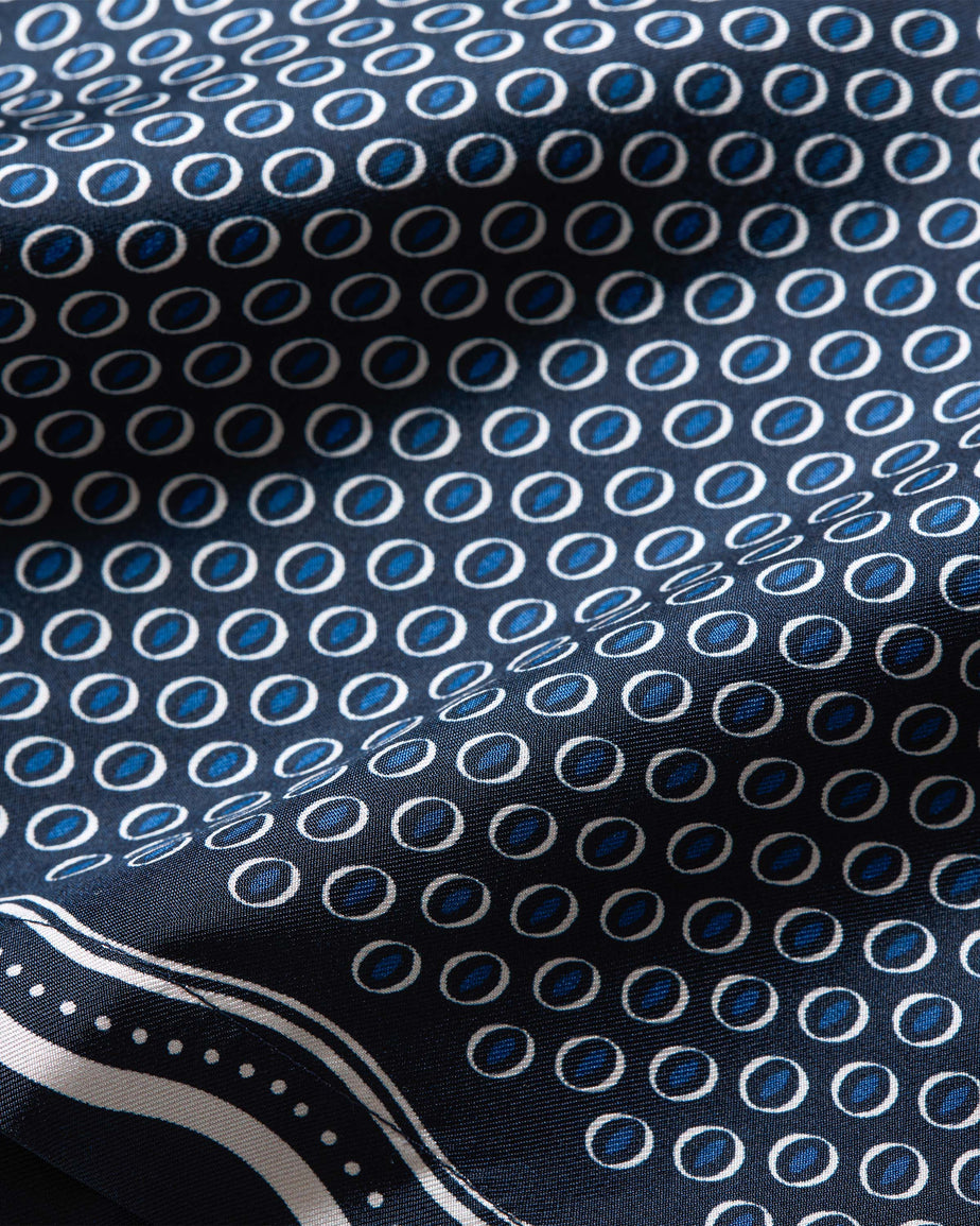 A close-up of a patterned fabric with a repeating motif of blue and white oval medallions on a dark navy background, with a decorative border visible in the lower left corner.
