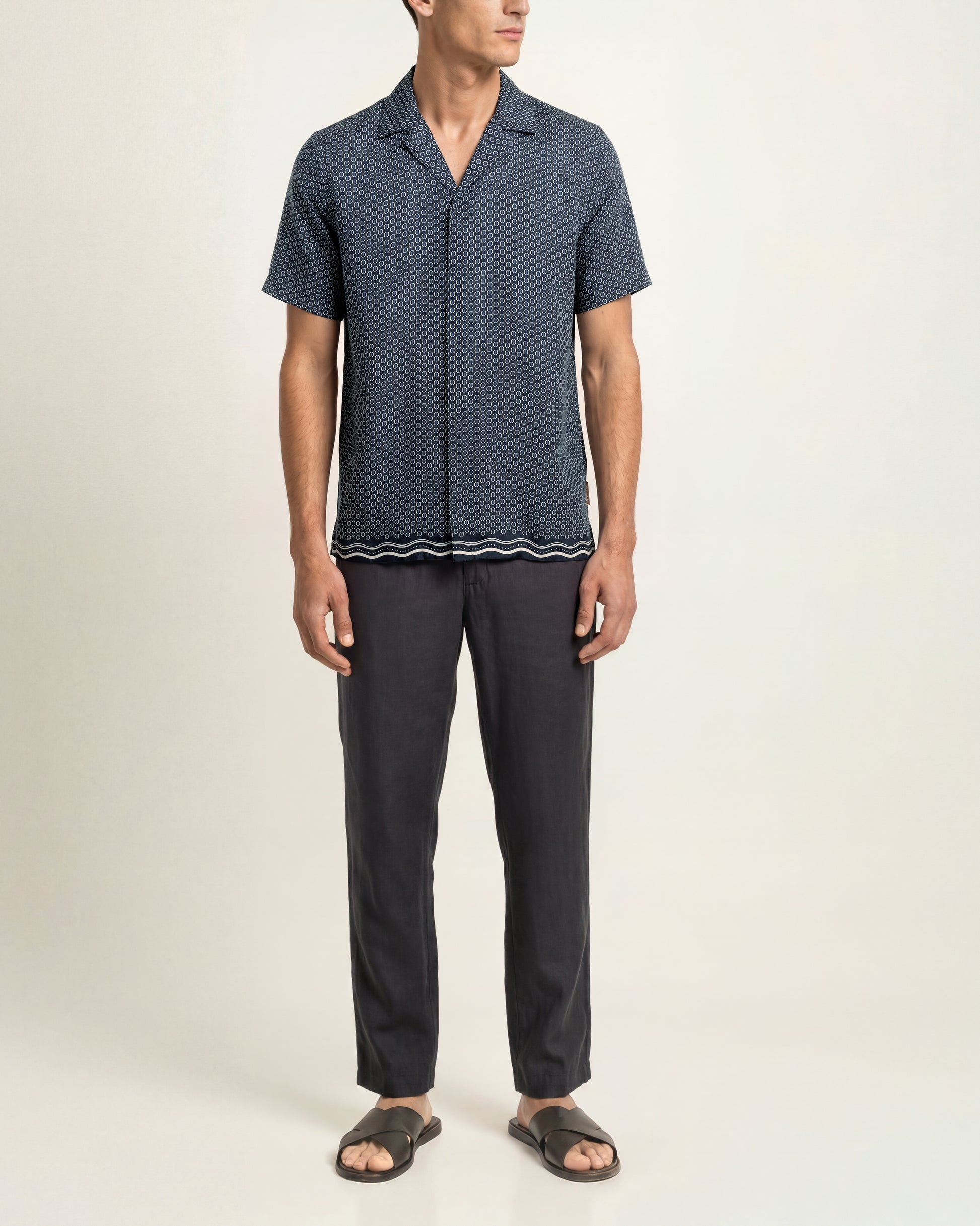 A man modeling a short-sleeve button-up shirt with a dark blue and black geometric pattern, paired with dark charcoal pants and black slide sandals, against a plain off-white studio background.