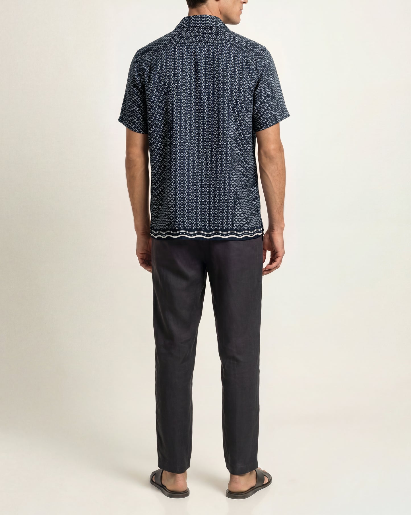 A man viewed from the back wearing a short-sleeve button-up shirt with a dark navy blue and black micro-pattern, paired with dark charcoal trousers and brown sandals, against a plain light beige studio background.