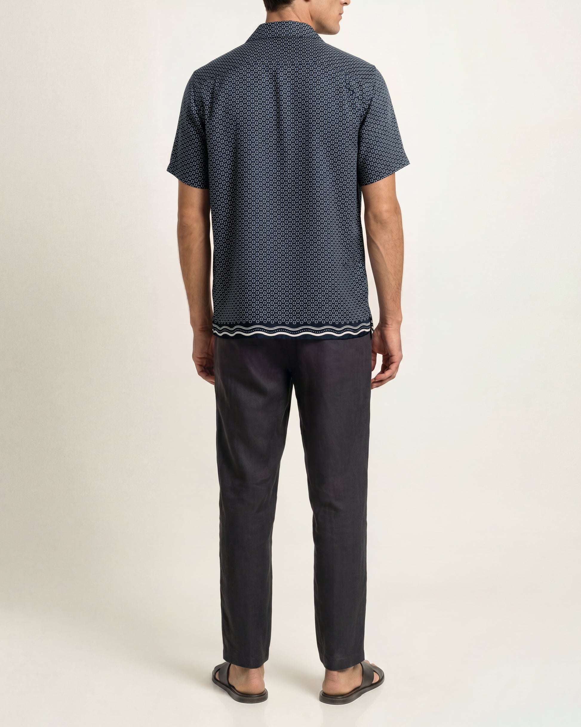A man viewed from the back wearing a short-sleeve button-up shirt with a dark navy blue and black micro-pattern, paired with dark charcoal trousers and brown sandals, against a plain light beige studio background.