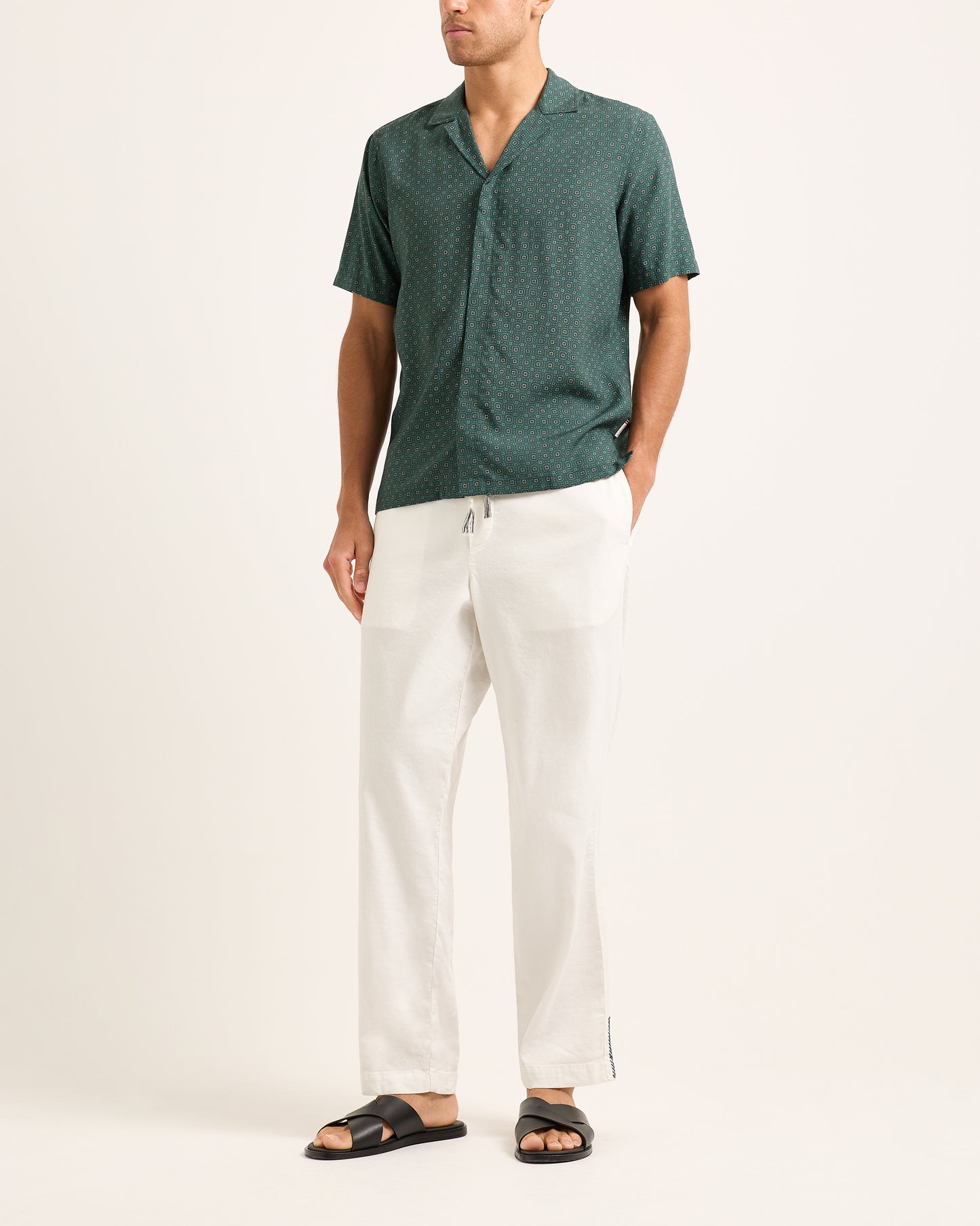 Model wearing dark green patterned short-sleeve shirt with white trousers on a white background
