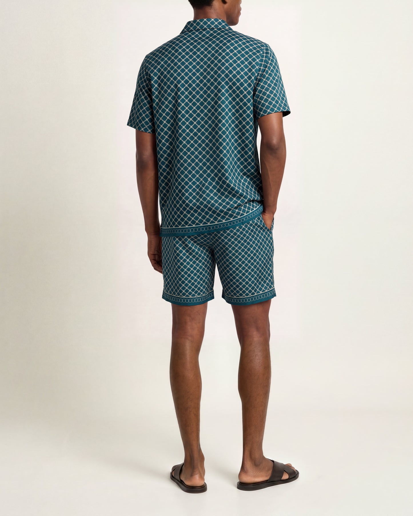 A man shown from the back wearing a matching short-sleeve collared shirt and shorts set in a dark teal green and white geometric pattern, paired with dark brown slide sandals, against a plain light beige studio background.