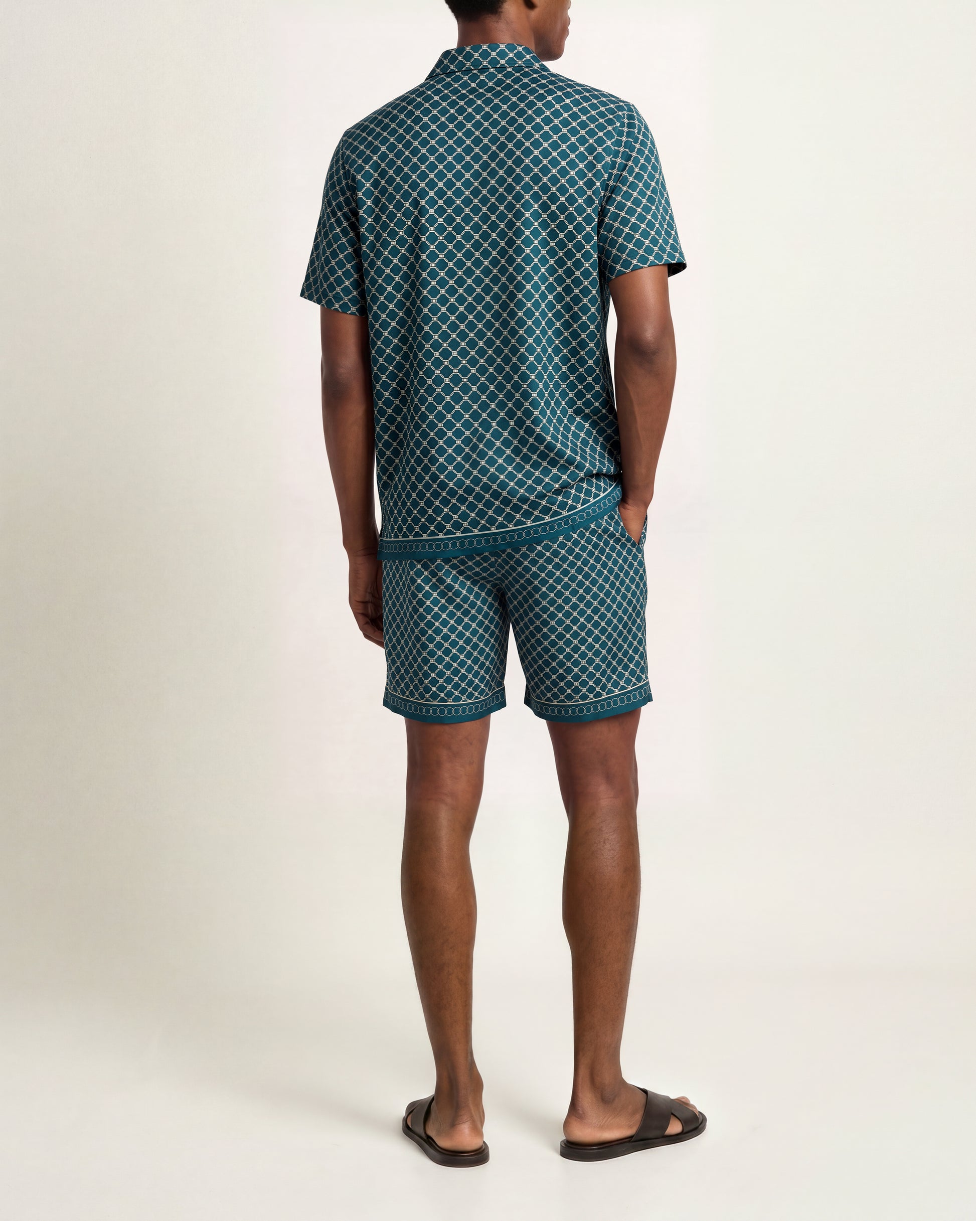 A man shown from the back wearing a matching short-sleeve collared shirt and shorts set in a dark teal green and white geometric pattern, paired with dark brown slide sandals, against a plain light beige studio background.