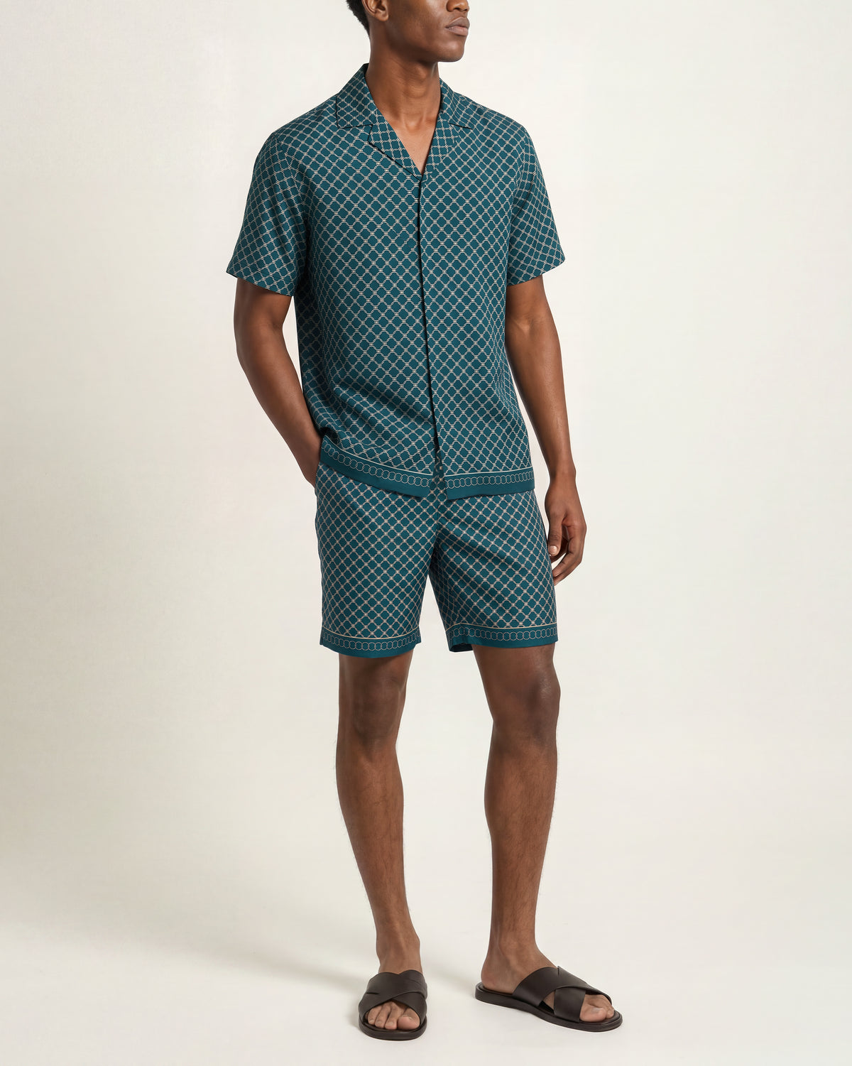 A man modeling a matching short-sleeve button-front shirt and shorts set in a dark teal green and white geometric pattern, paired with dark brown slide sandals, against a plain off-white studio background.