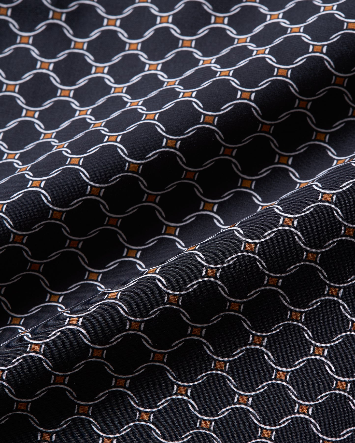 A close-up of a woven fabric with a repeating geometric pattern of interlocking chain links in white, accented by small orange diamond shapes on a very dark navy or black background.