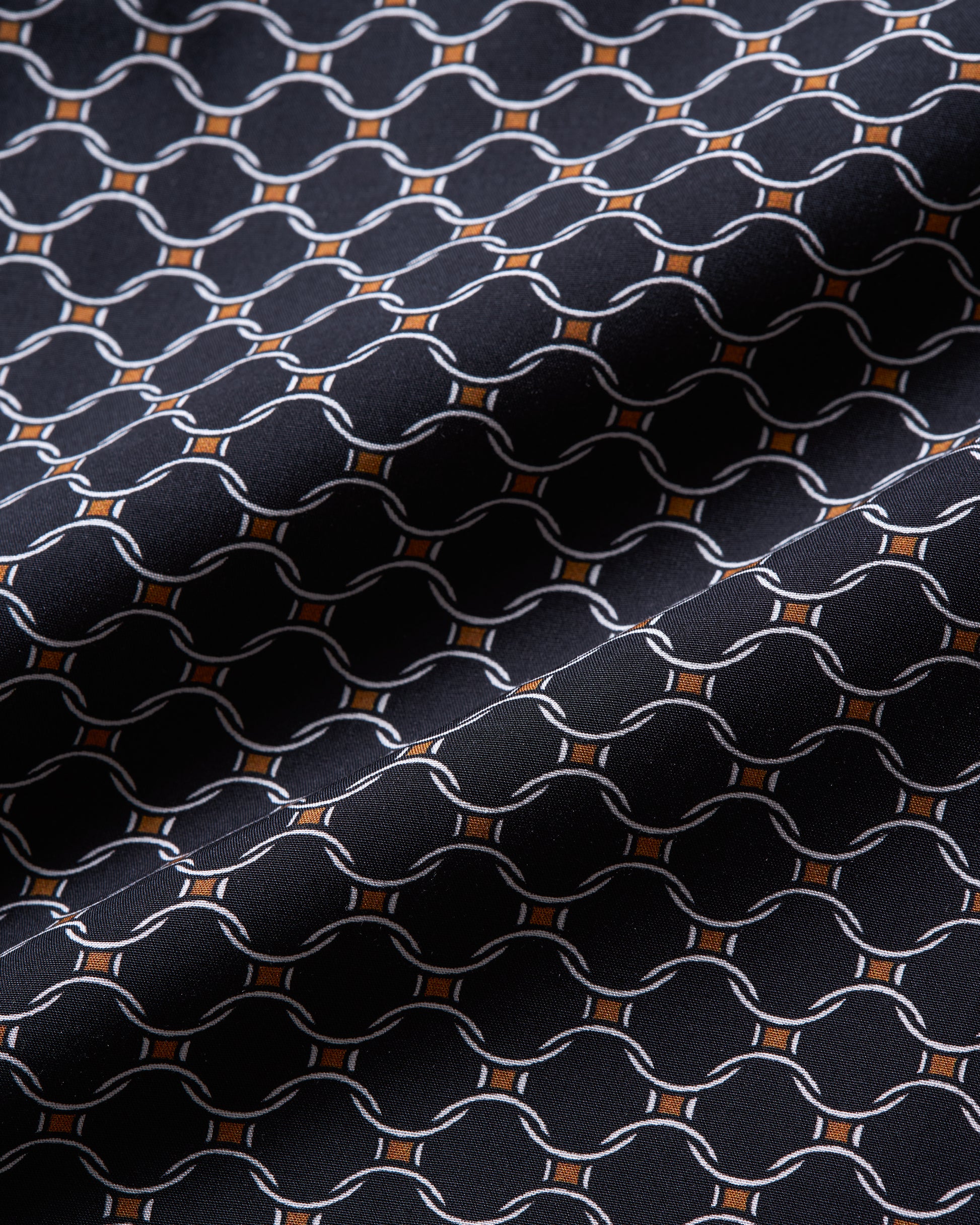 A close-up of a woven fabric with a repeating geometric pattern of interlocking chain links in white, accented by small orange diamond shapes on a very dark navy or black background.