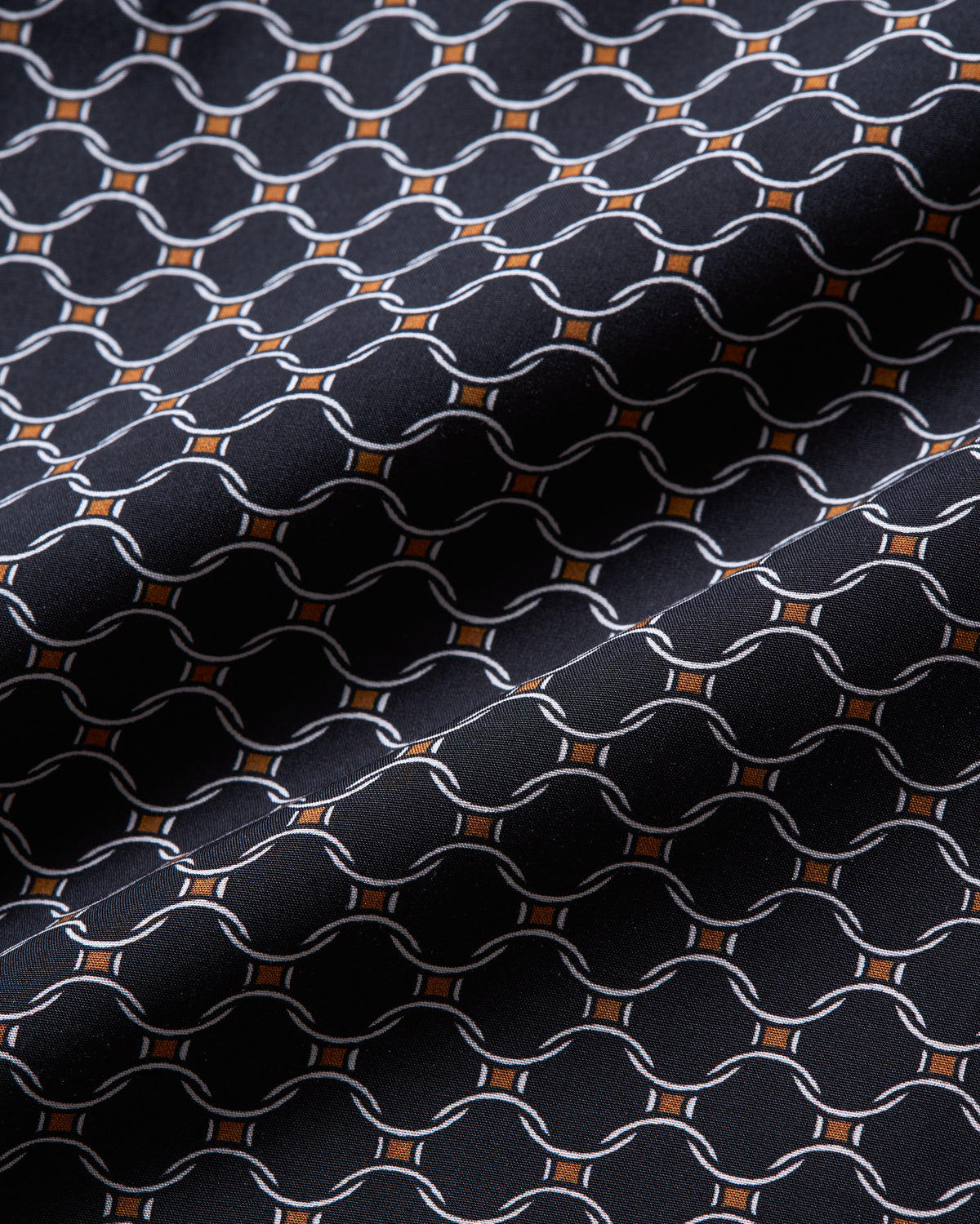 A close-up of a woven fabric with a repeating geometric pattern of interlocking chain links in white, accented by small orange diamond shapes on a very dark navy or black background.