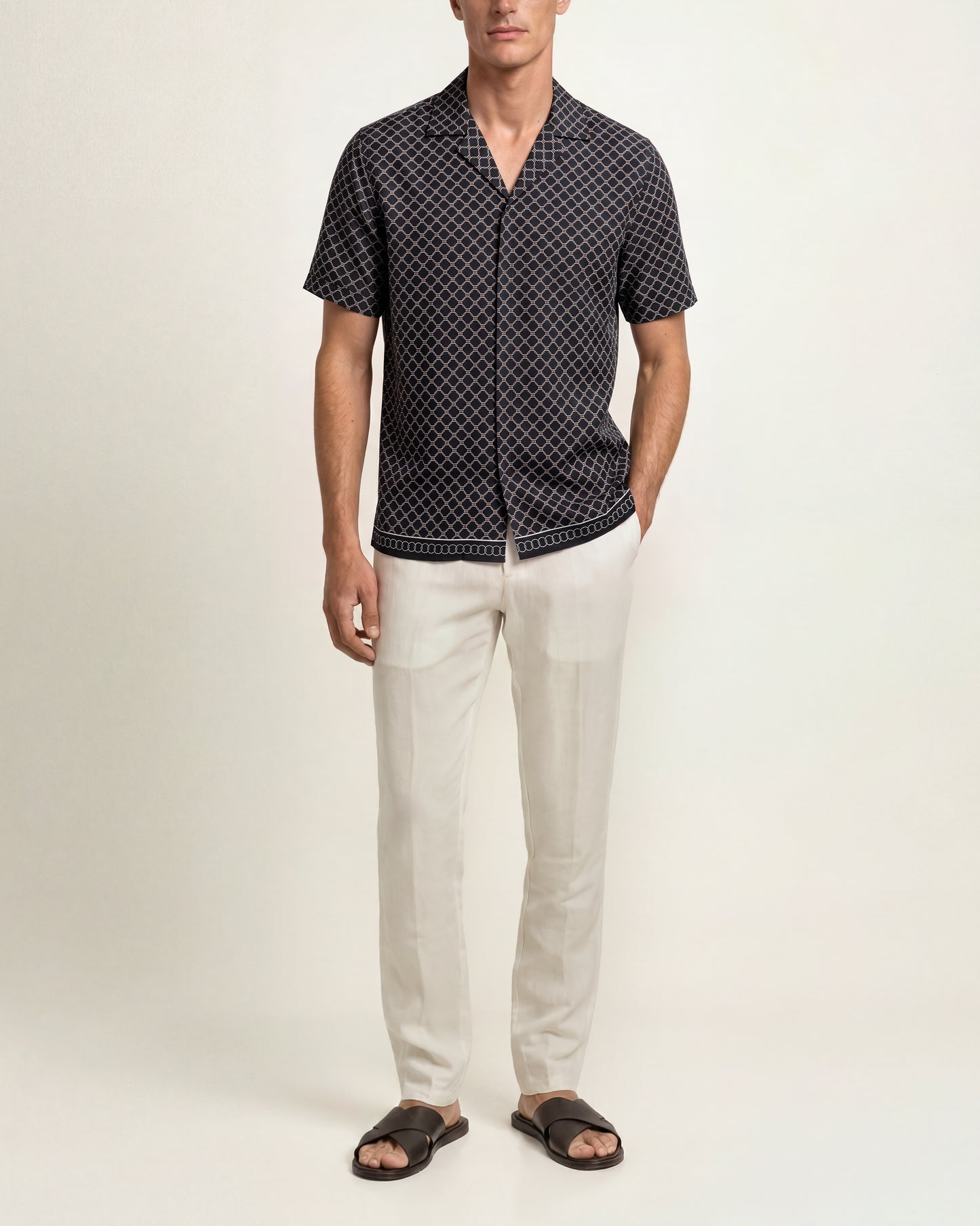 A man modeling a short-sleeve button-up shirt with a black and white geometric pattern, paired with off-white trousers and dark brown slide sandals, against a plain light beige studio background.