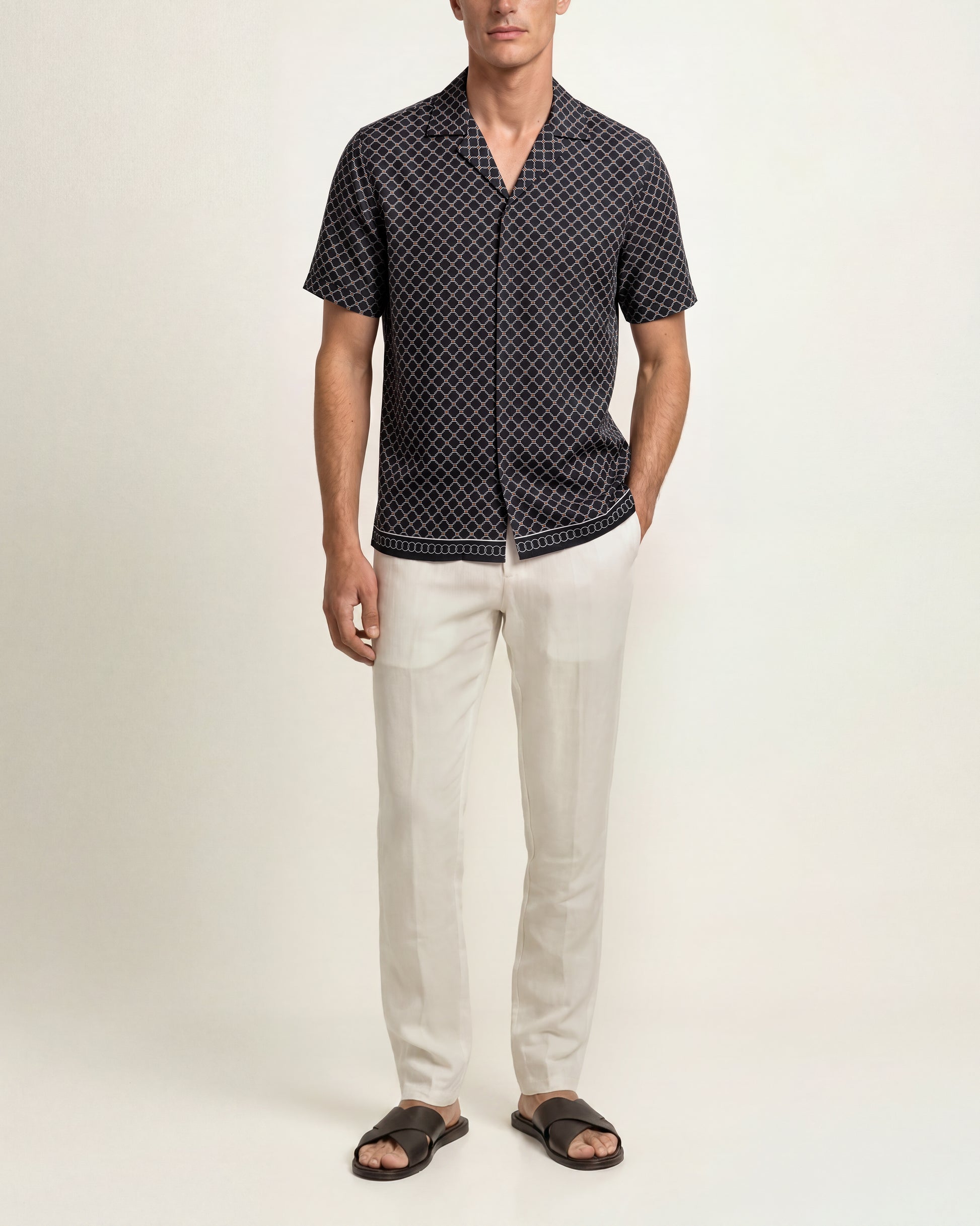 A man modeling a short-sleeve button-up shirt with a black and white geometric pattern, paired with off-white trousers and dark brown slide sandals, against a plain light beige studio background.