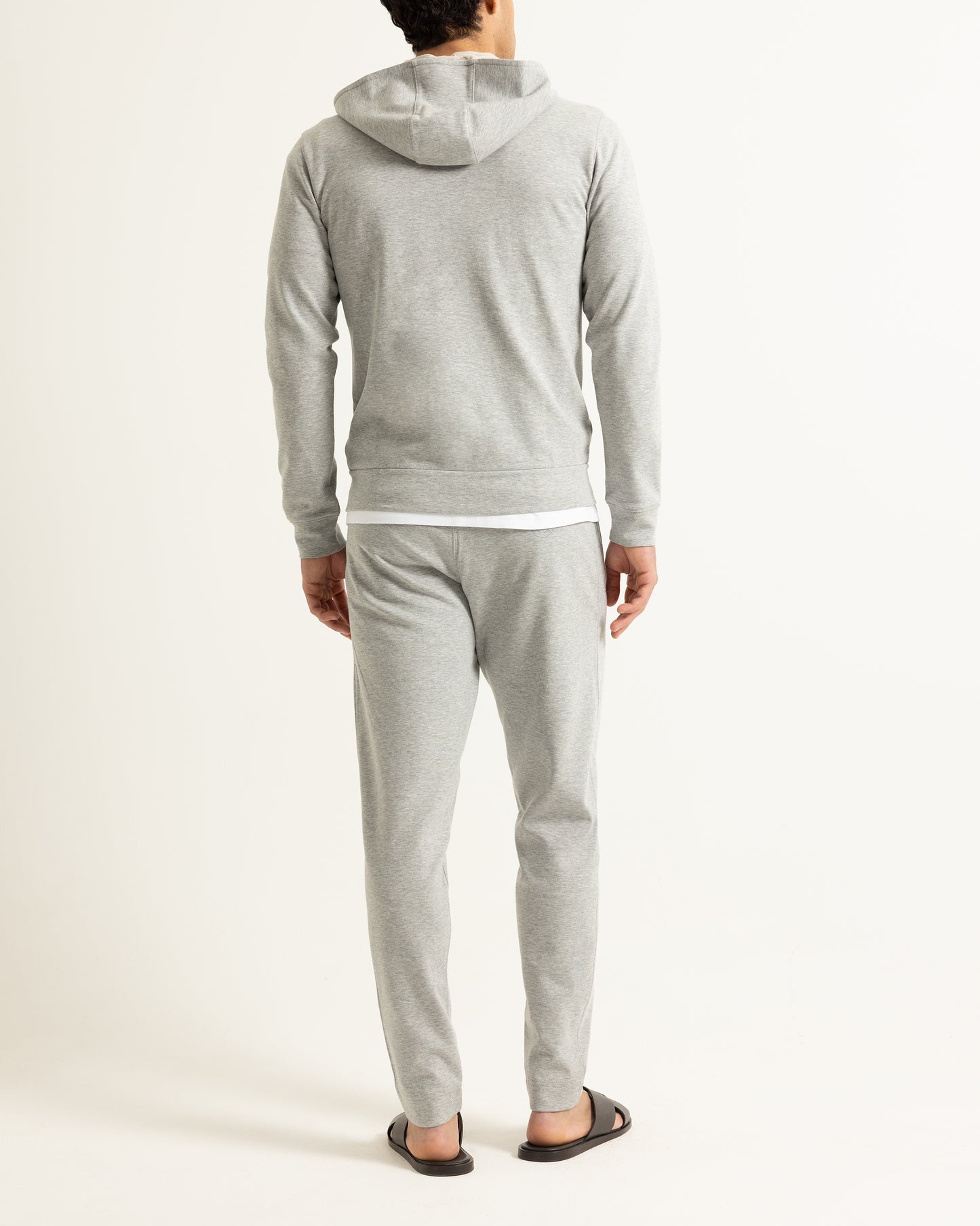 A man viewed from behind wearing a light gray heathered hooded sweatshirt and matching jogger pants, with black sandals, against a plain off-white studio background.