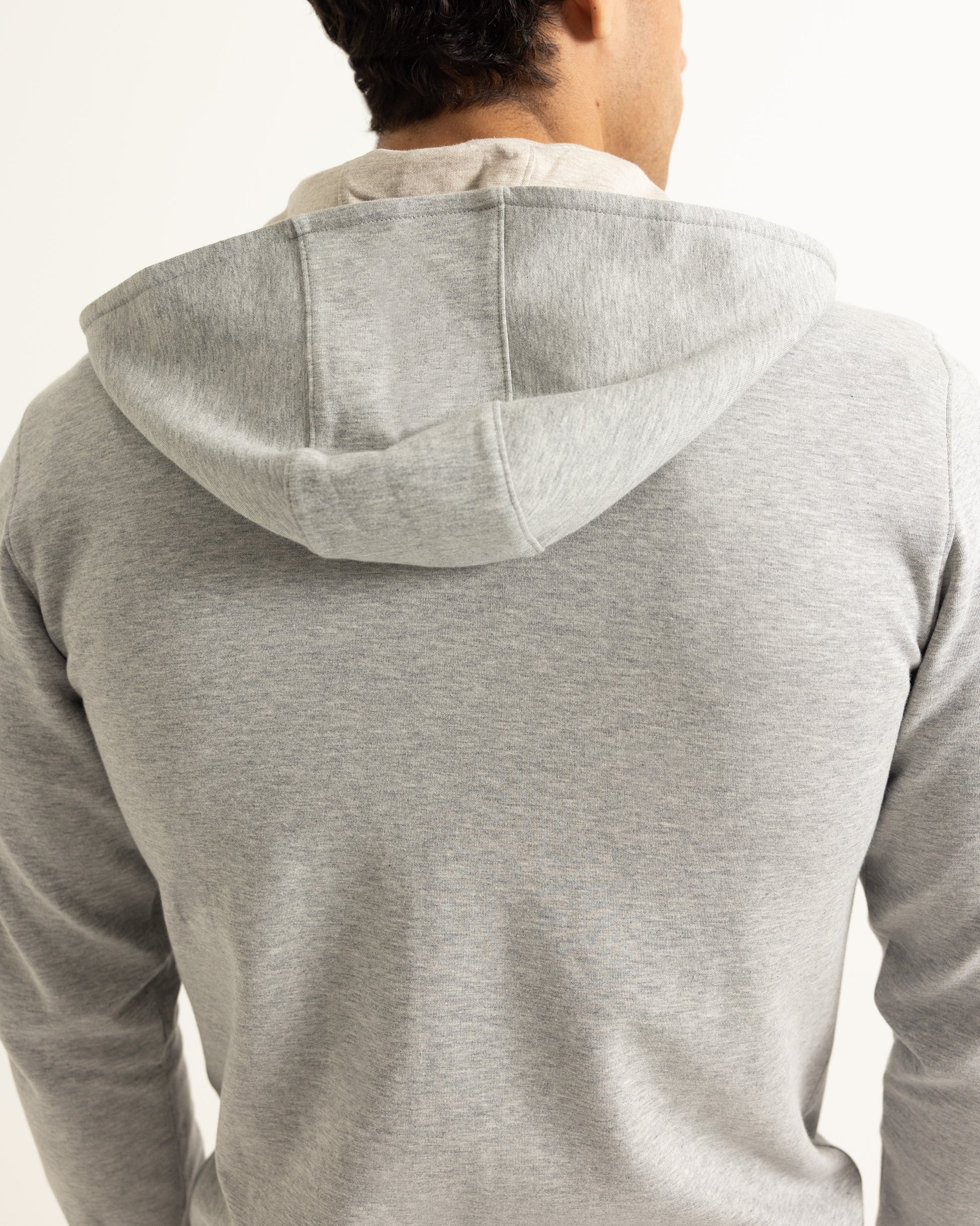 A man viewed from behind wearing a light gray heathered hoodie with a beige undershirt, against a plain white background.