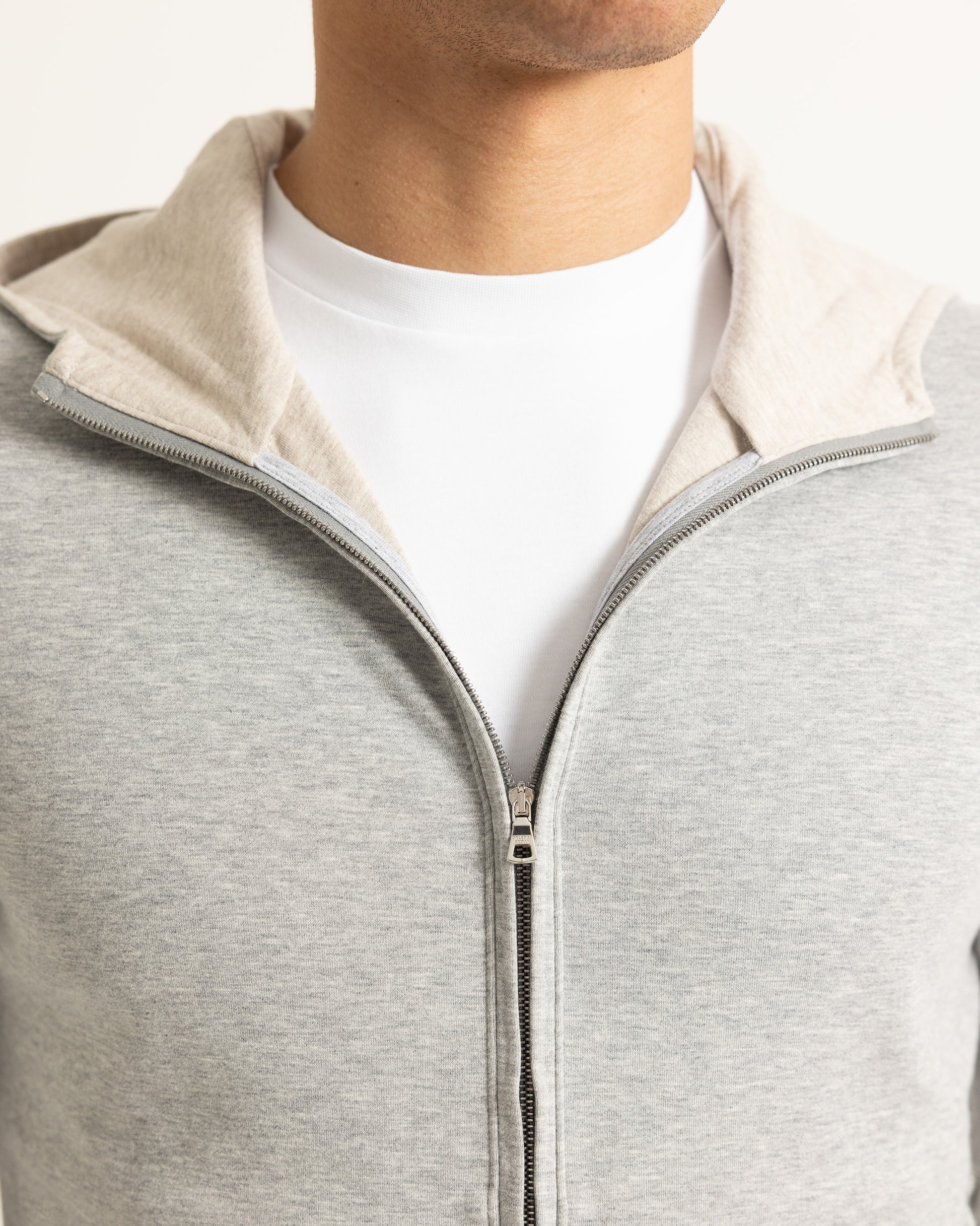 Close-up of a person wearing a light gray heather zip-up hoodie with a beige inner collar, layered over a white crew-neck t-shirt.