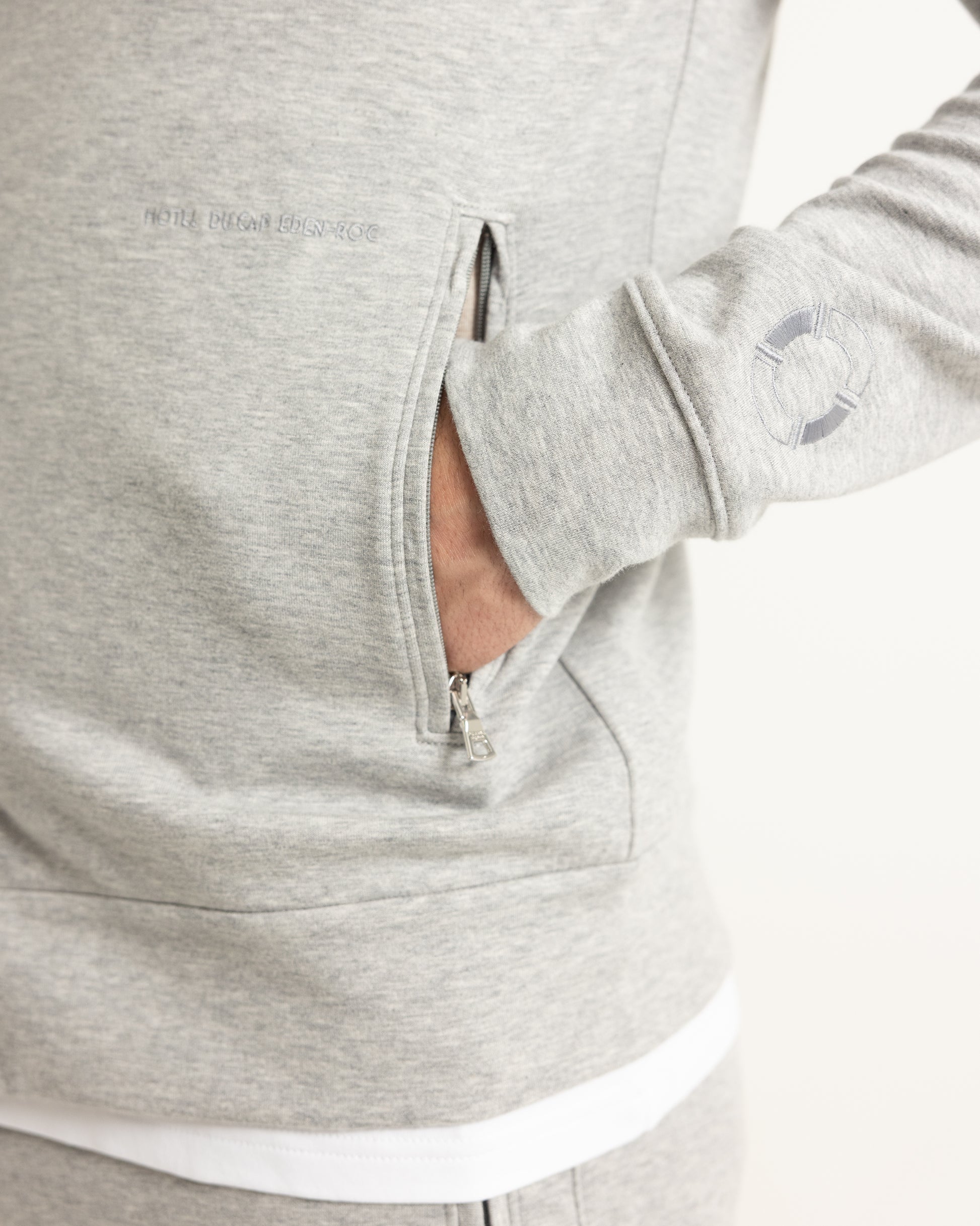 Close-up of a person wearing a light heather gray hoodie, showing a side zip pocket with a hand inside and a small circular logo on the sleeve.