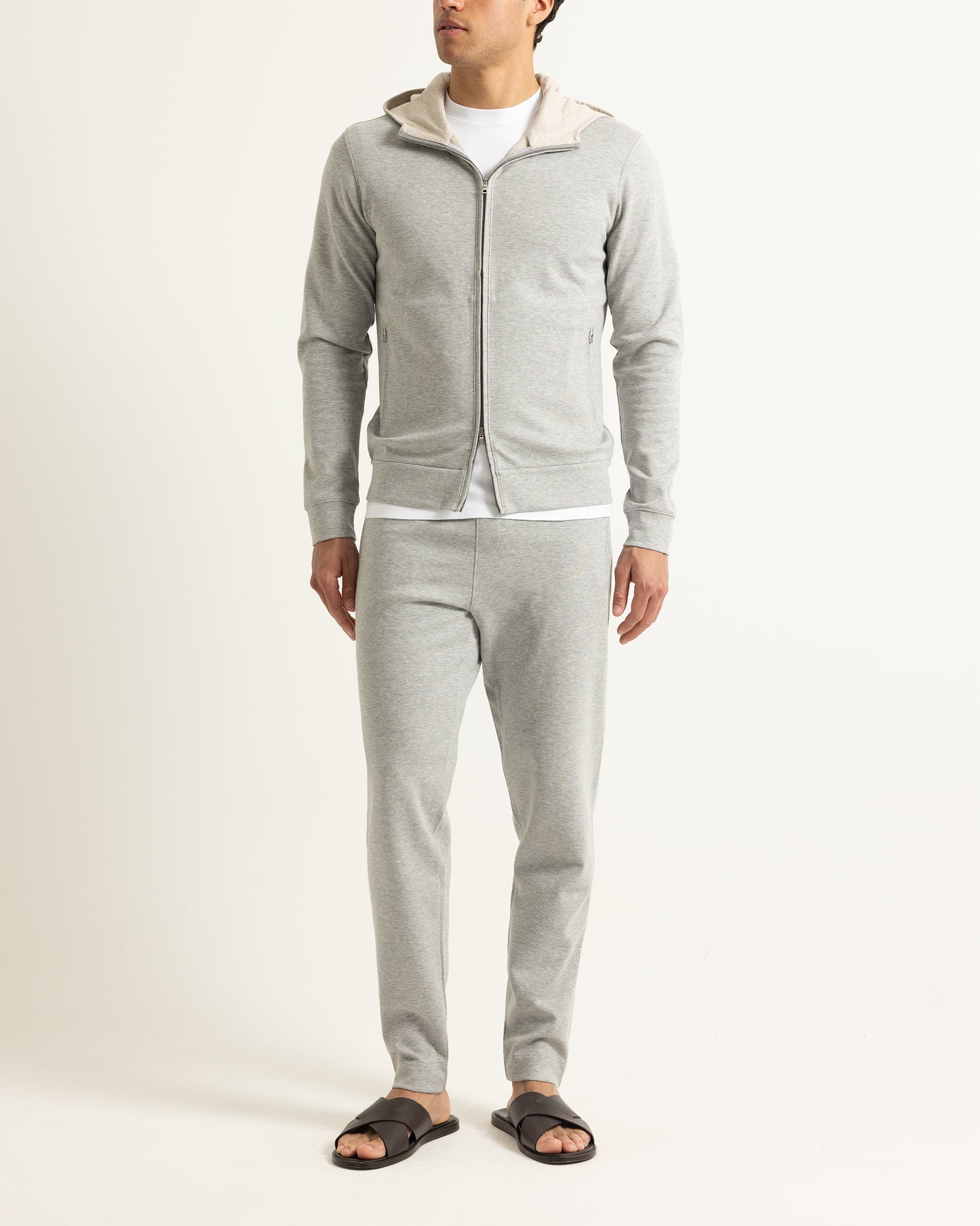 A man modeling a light gray zip-up hoodie with a beige hood lining and matching light gray sweatpants, styled with a white t-shirt and black slide sandals against a plain off-white studio background.