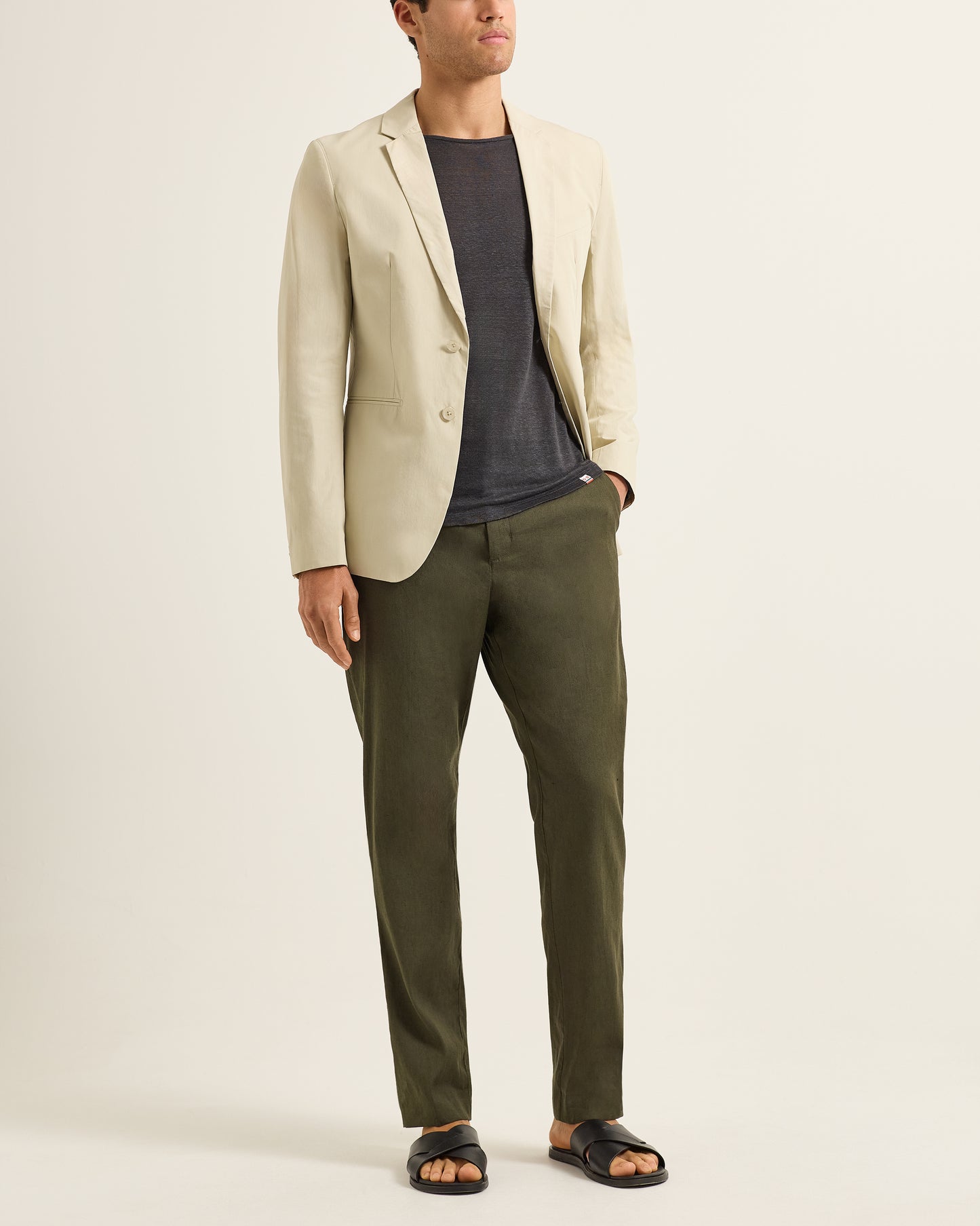 Man wearing a beige blazer, dark shirt, and green pants on a plain background