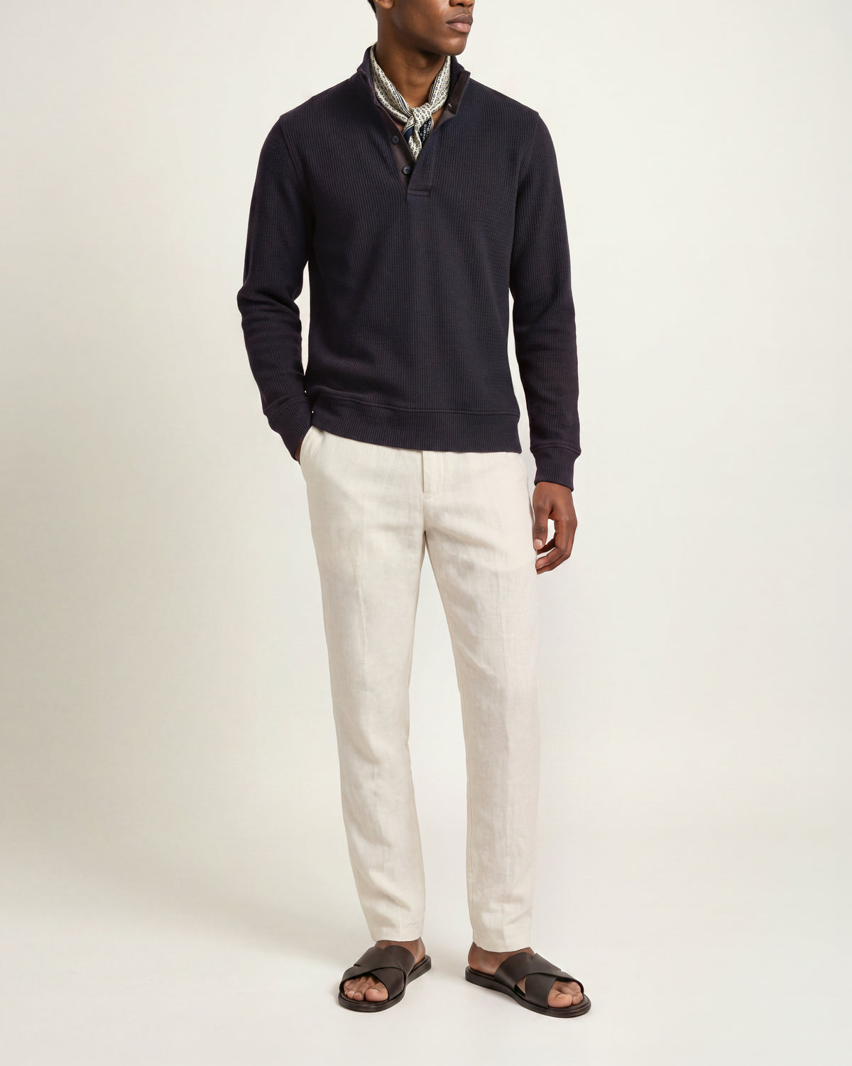 Man wearing navy long sleeve sweatshirt with white trousers and brown sandals on  a plain background
