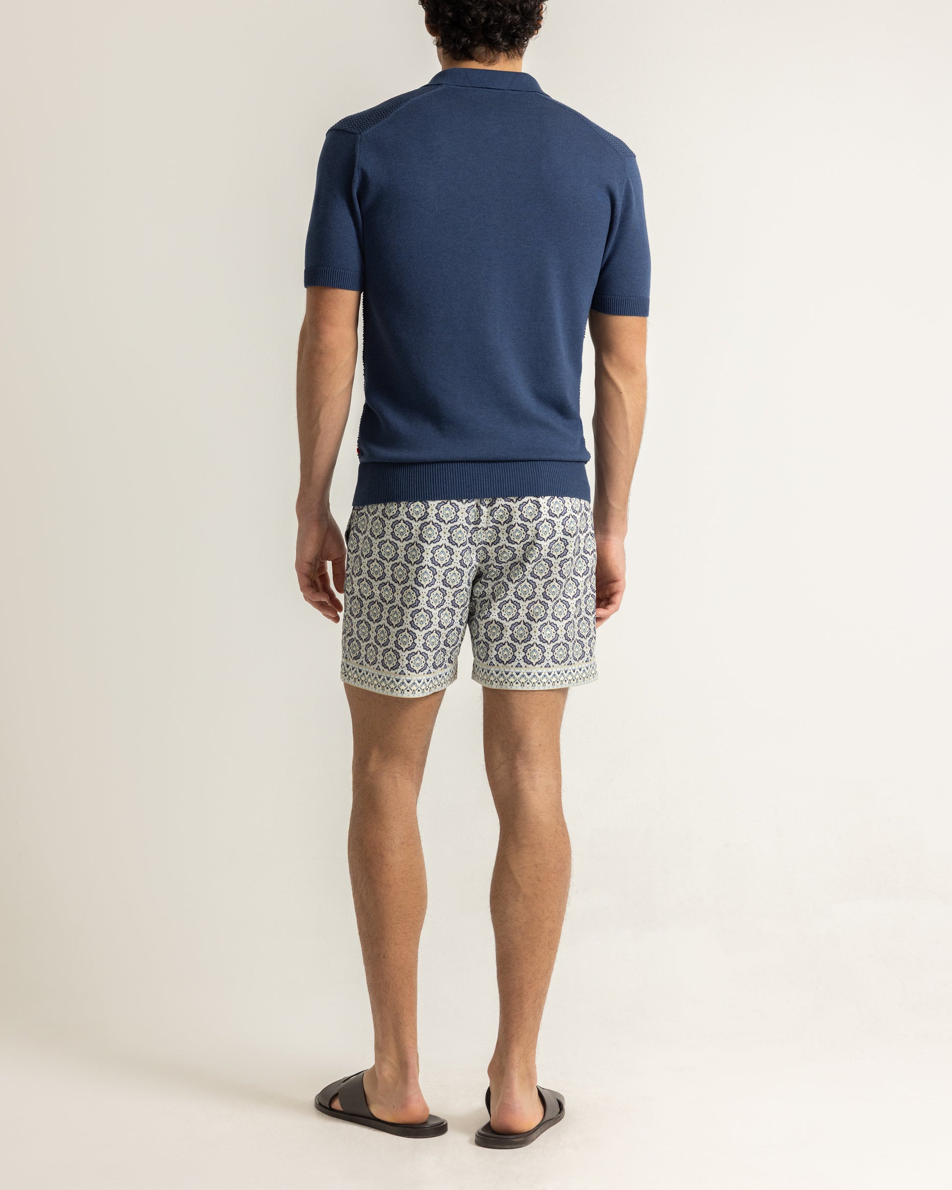 Man wearing a navy polo shirt with patterned swim shorts on a plain background