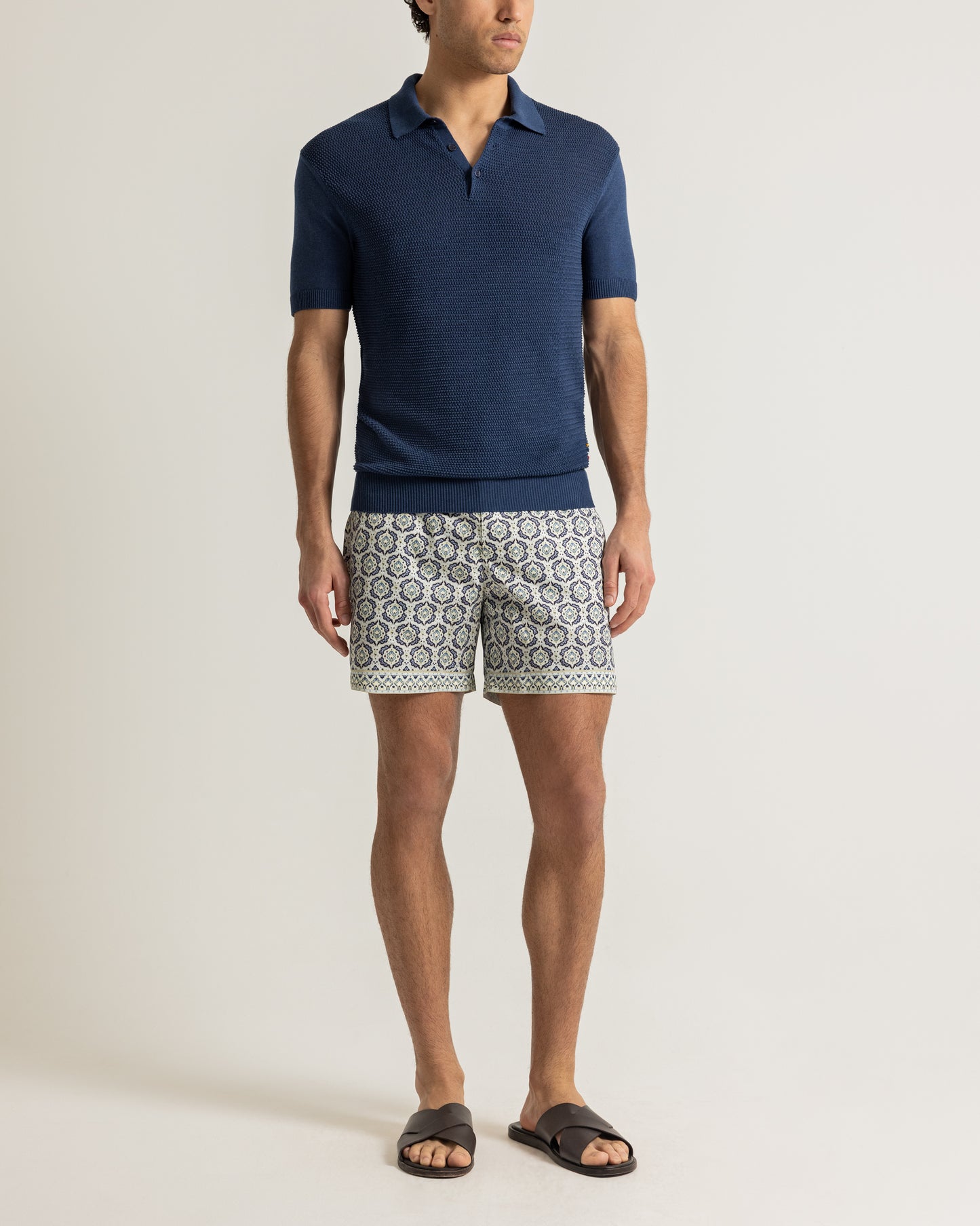 Man wearing a navy polo shirt with patterned swim shorts on a plain background