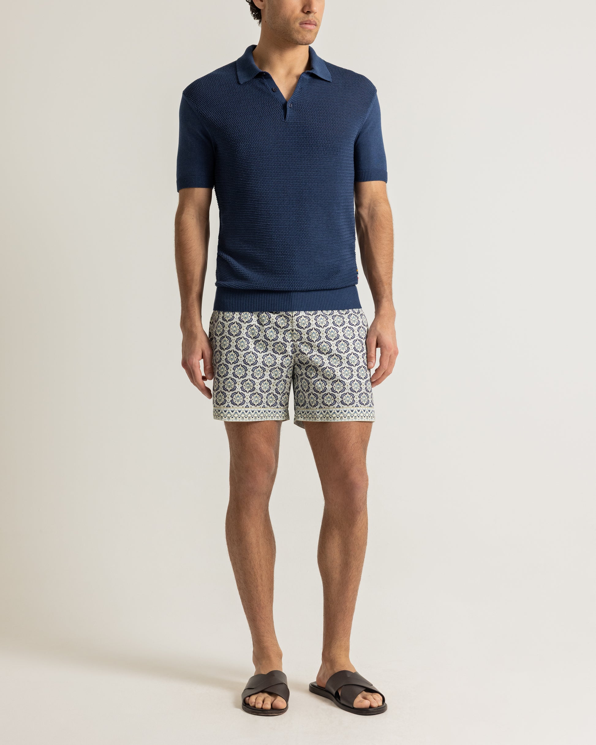 Man wearing a navy polo shirt with patterned swim shorts on a plain background