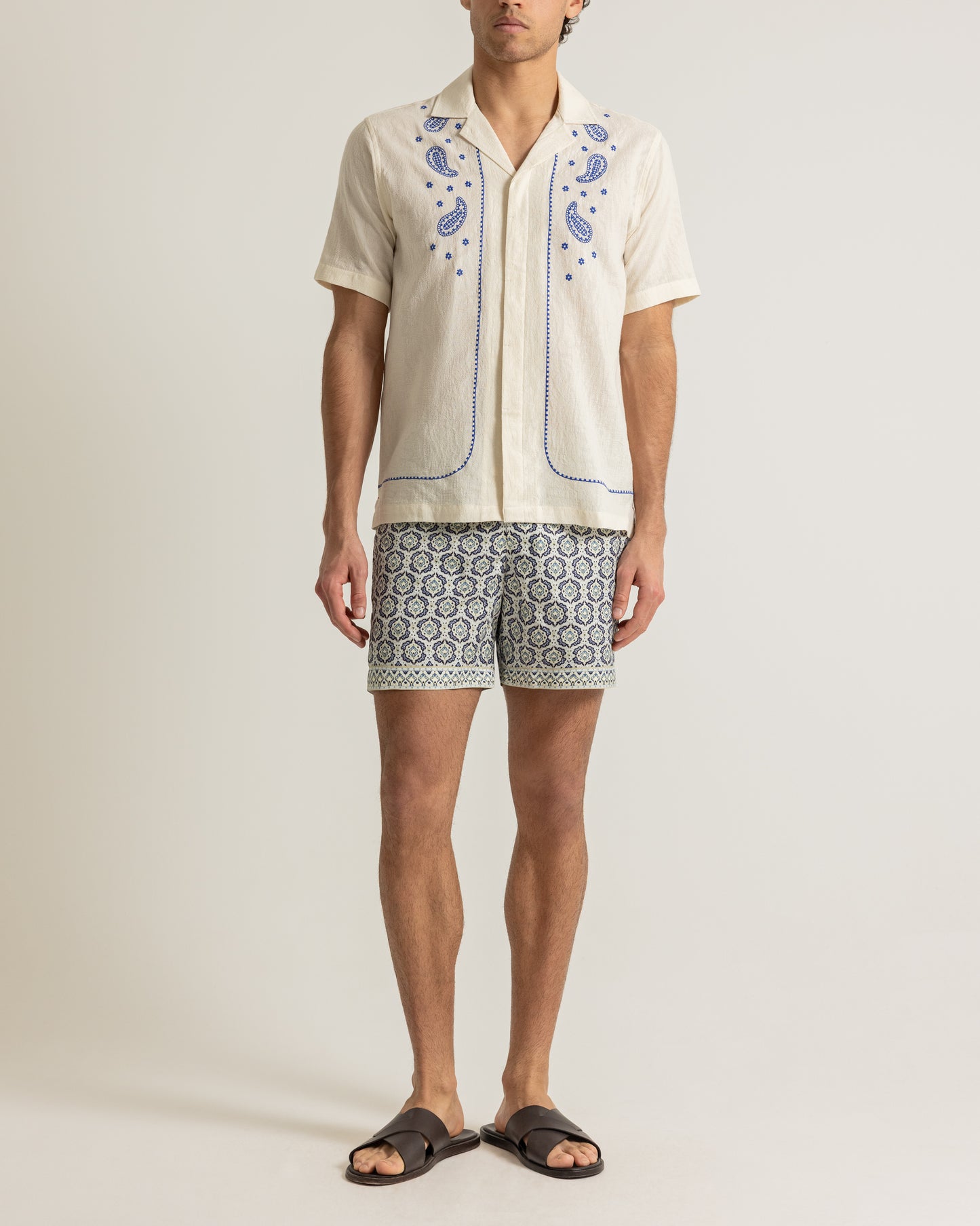 Man wearing white printed white shirt with patterned swim shorts on a plain background 