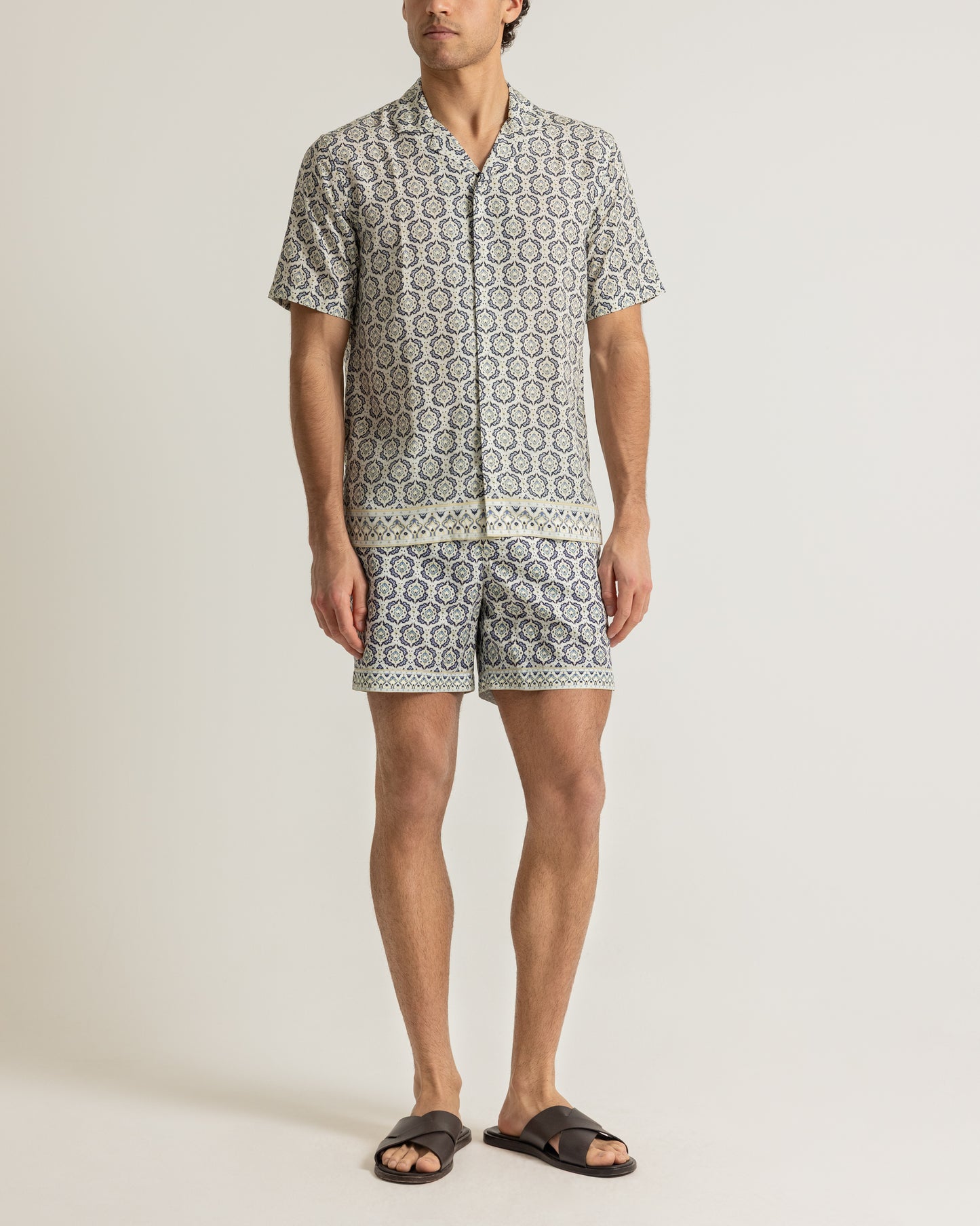 Man wearing printed beige shirt and swim shorts on a plain background