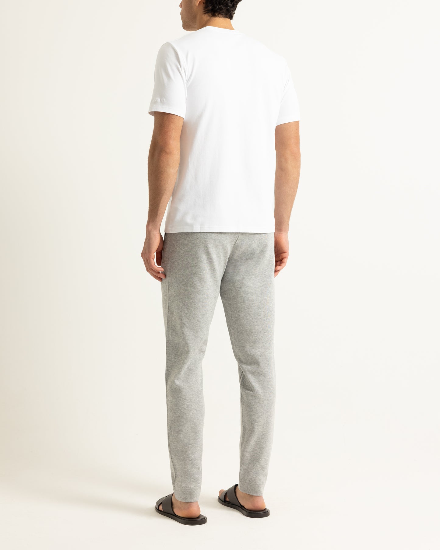 A man viewed from behind wearing a white short-sleeve t-shirt, light gray heathered sweatpants, and black slide sandals, standing against a plain off-white studio background.