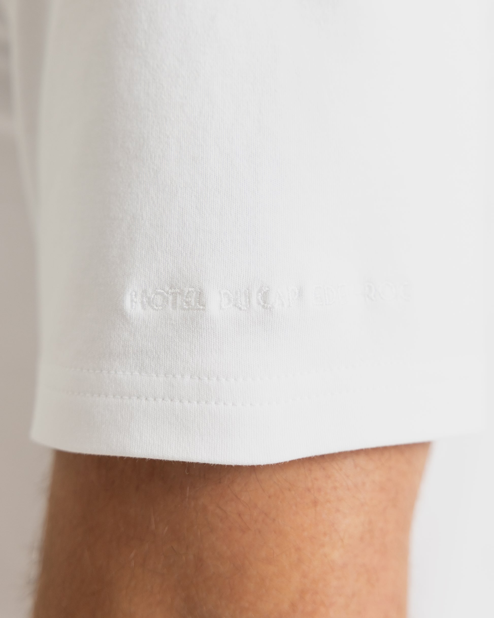 Close-up of a white short-sleeve t-shirt sleeve with a ribbed hem and subtle tonal embroidered text on the cuff, worn on a person's arm.