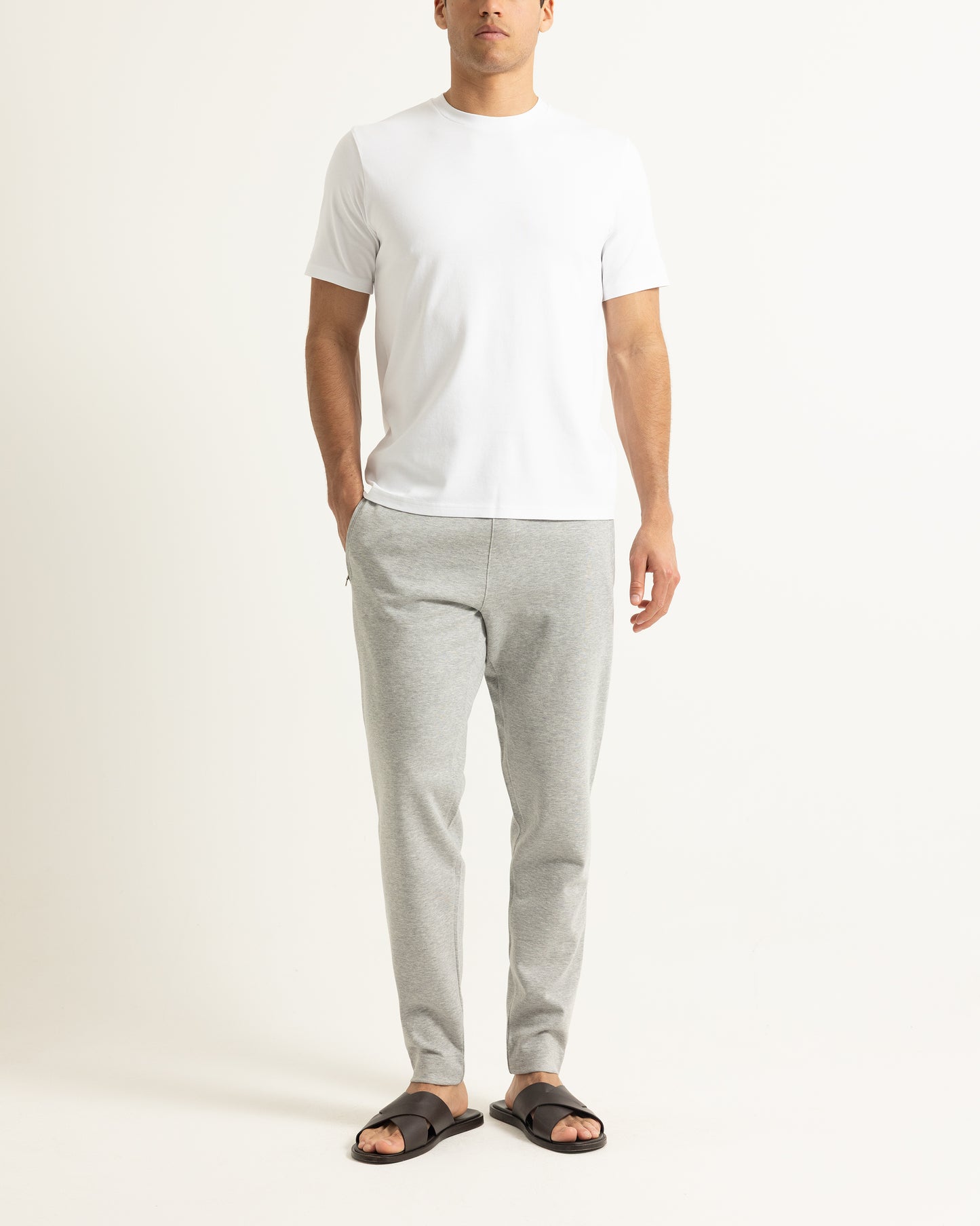 A man wearing a plain white short-sleeve crewneck t-shirt, light gray heathered sweatpants, and black slide sandals, standing against a solid off-white studio background.
