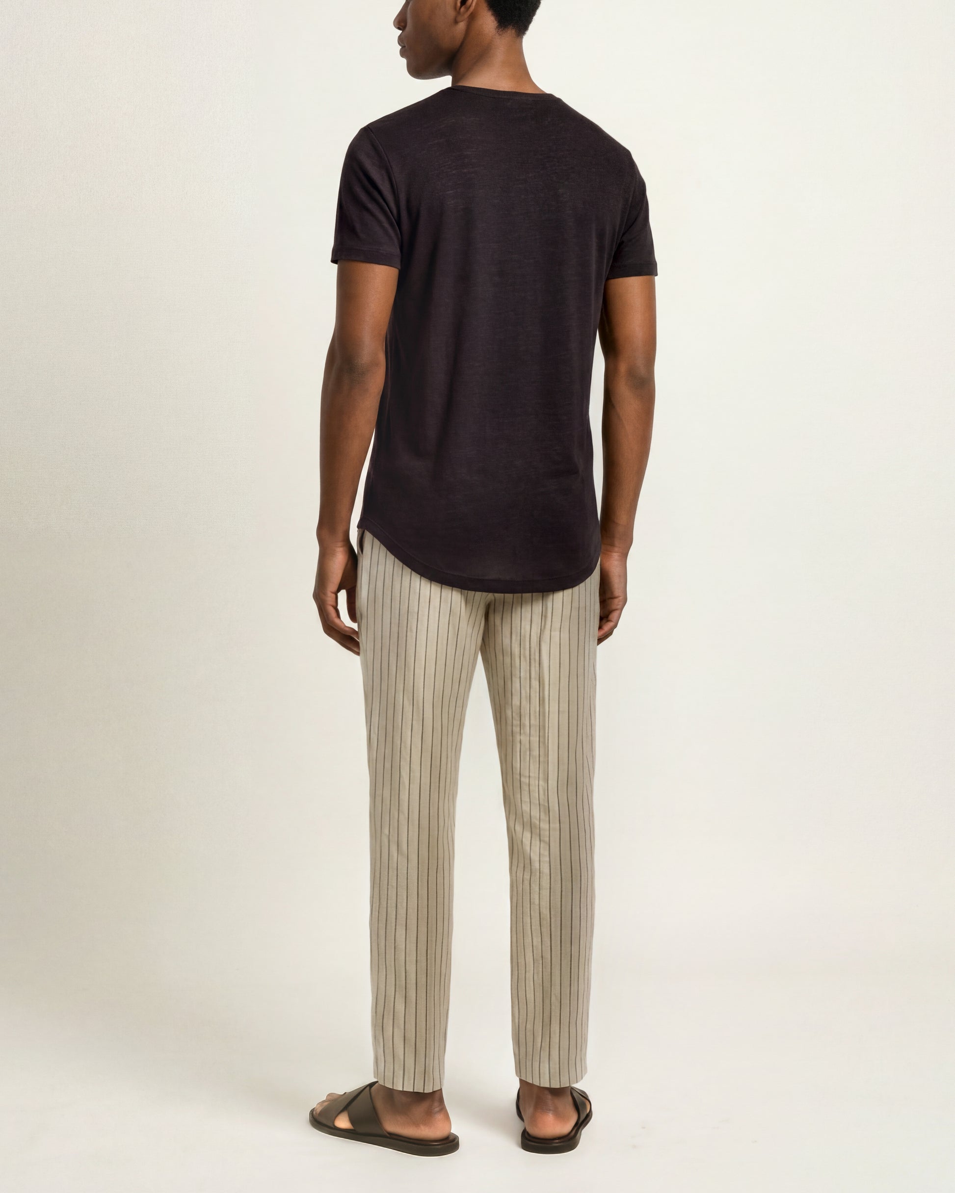 A man shown from the back wearing a dark charcoal short-sleeve t-shirt, beige and brown vertically striped trousers, and dark brown slide sandals, against a plain off-white studio background.