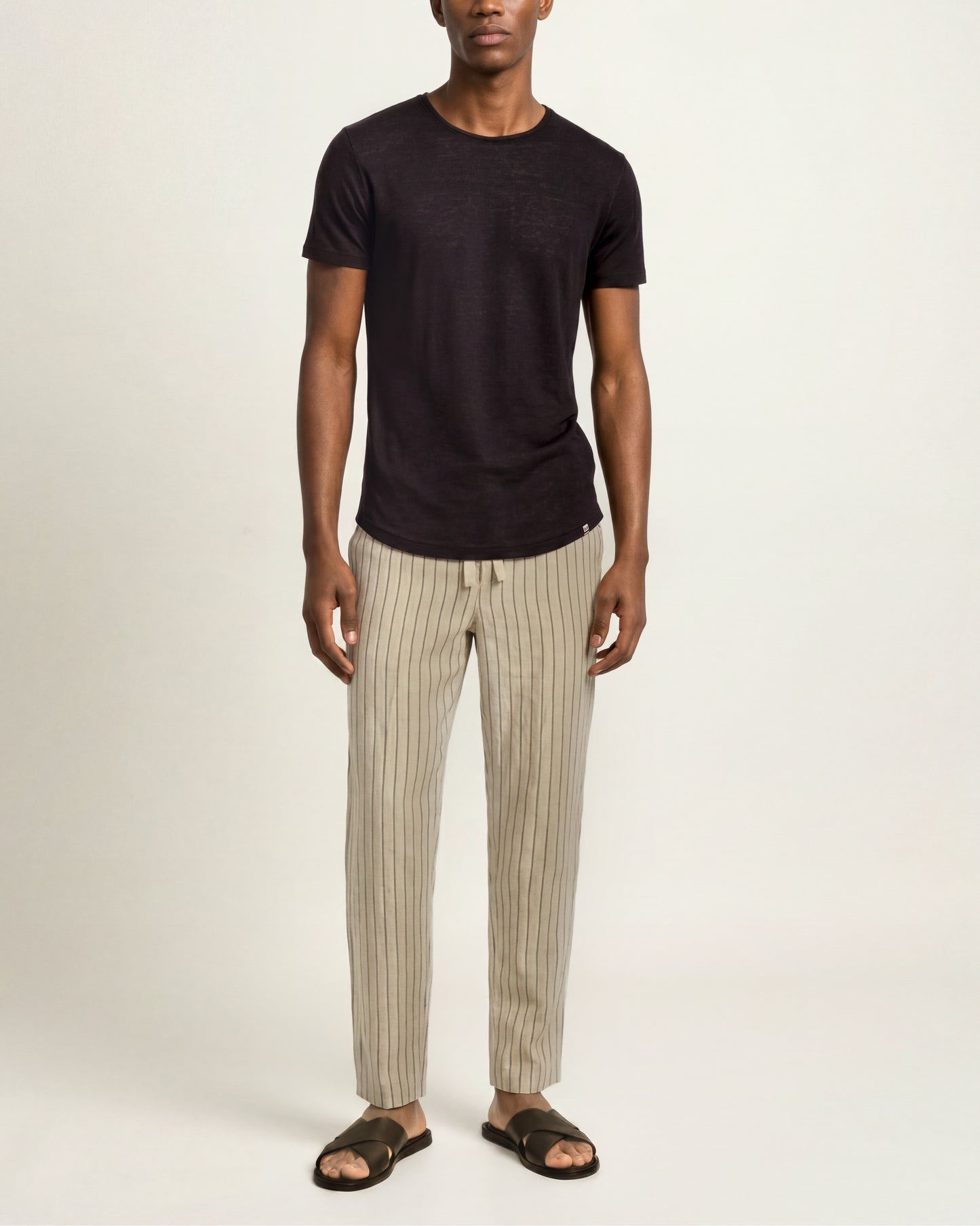 A man modeling a dark charcoal t-shirt and beige pinstriped trousers with dark brown slide sandals, standing against a plain off-white studio background.