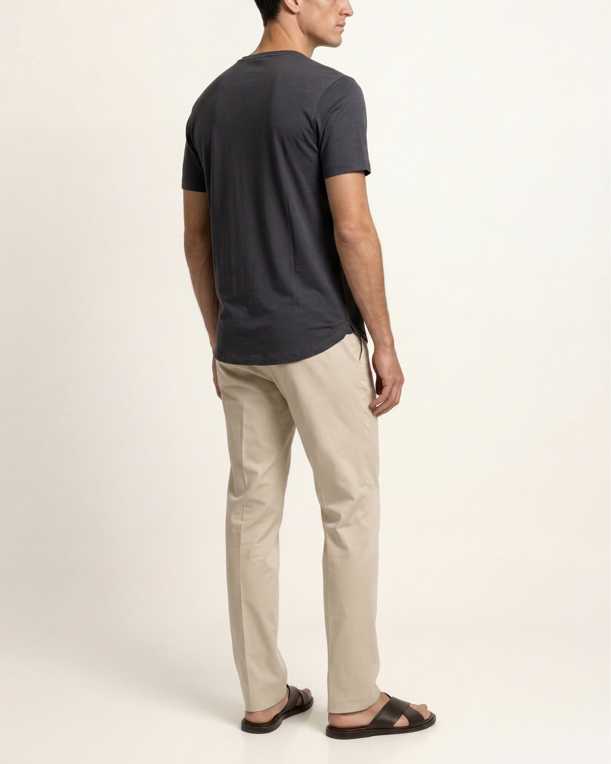 A man shown from the back wearing a dark charcoal gray t-shirt, beige straight-leg trousers, and dark brown sandals, photographed against a clean off-white studio background.