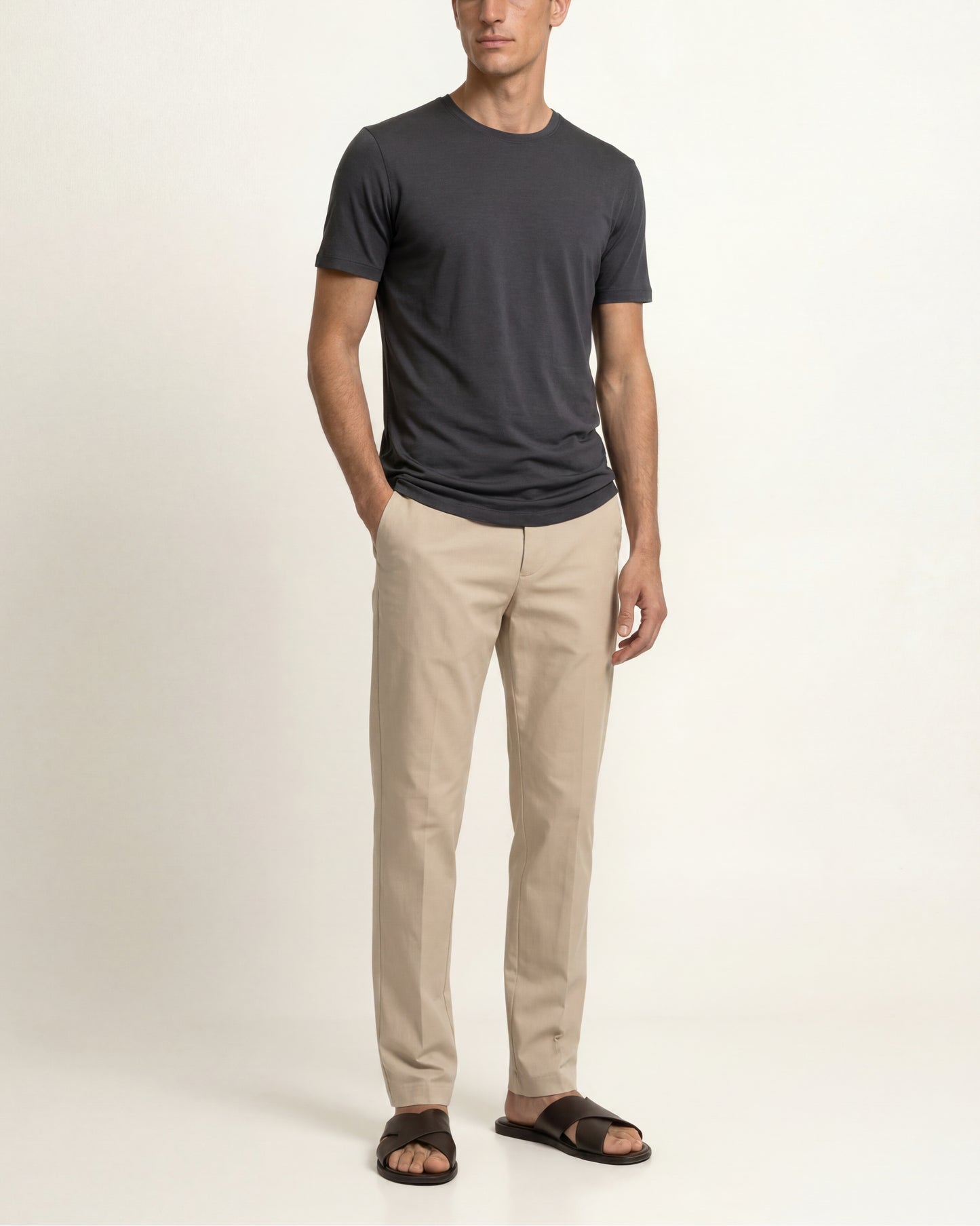 A man modeling a dark charcoal gray crewneck t-shirt, beige straight-leg chinos, and dark brown slide sandals against a plain off-white studio background.