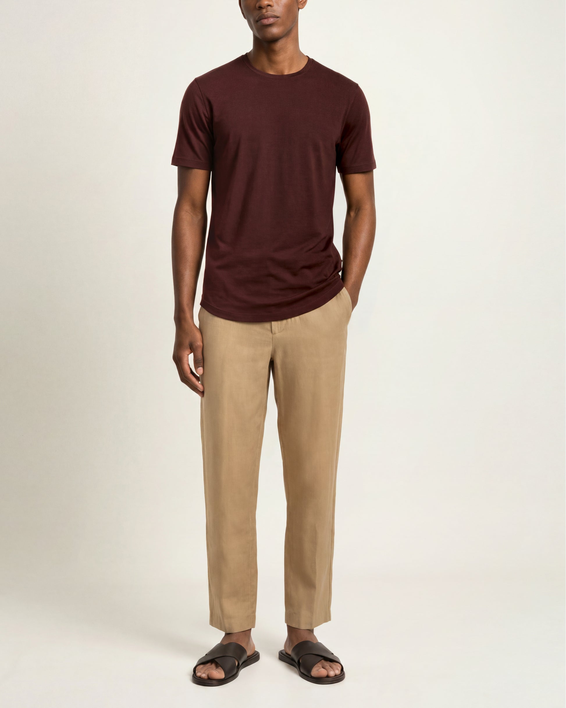 A man modeling a dark brown short-sleeve crewneck t-shirt, beige straight-leg trousers, and dark brown slide sandals against a plain off-white studio background.